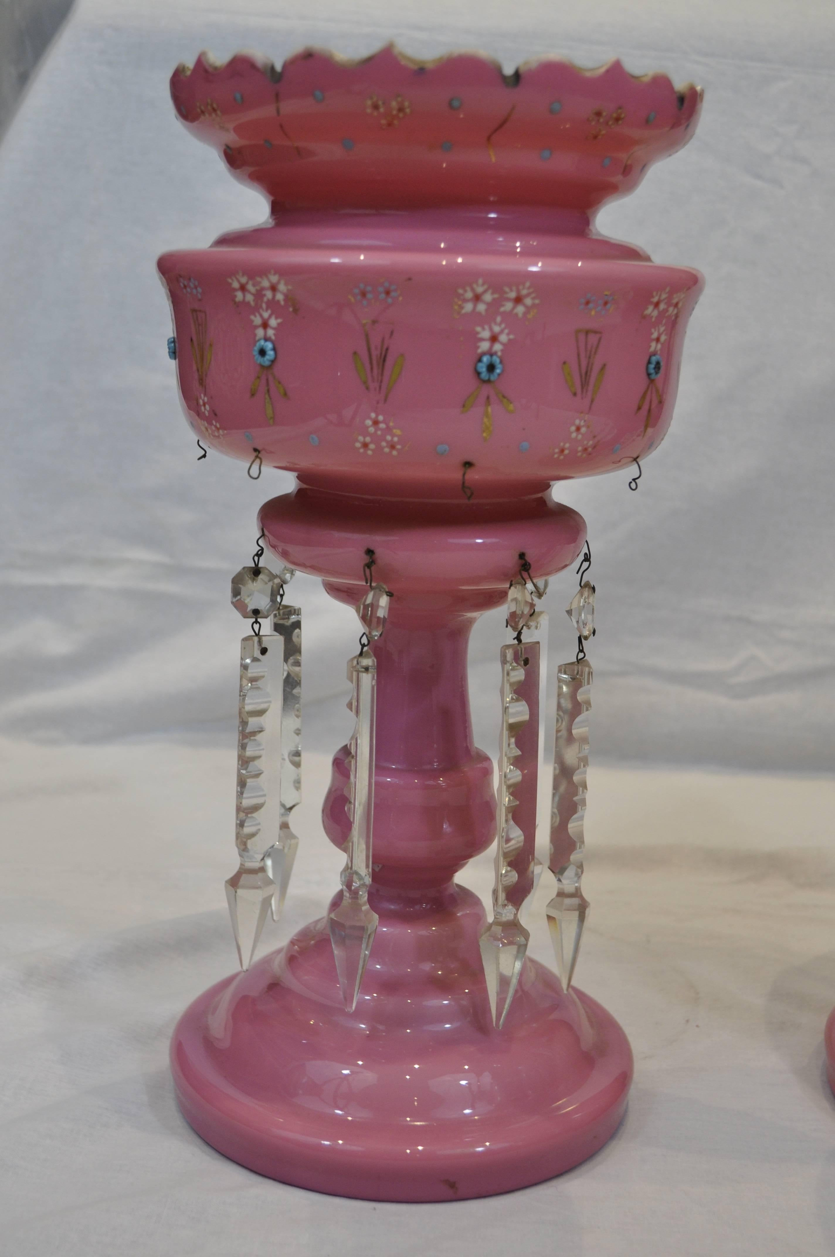 A pair of pink Bohemian opaline glass mantel lustres in the shape of a large cup with a scalloped rim with a round stand and base. They are decorated with white flower enameled designs and feature small blue glass ‘jewels’ that form further floral