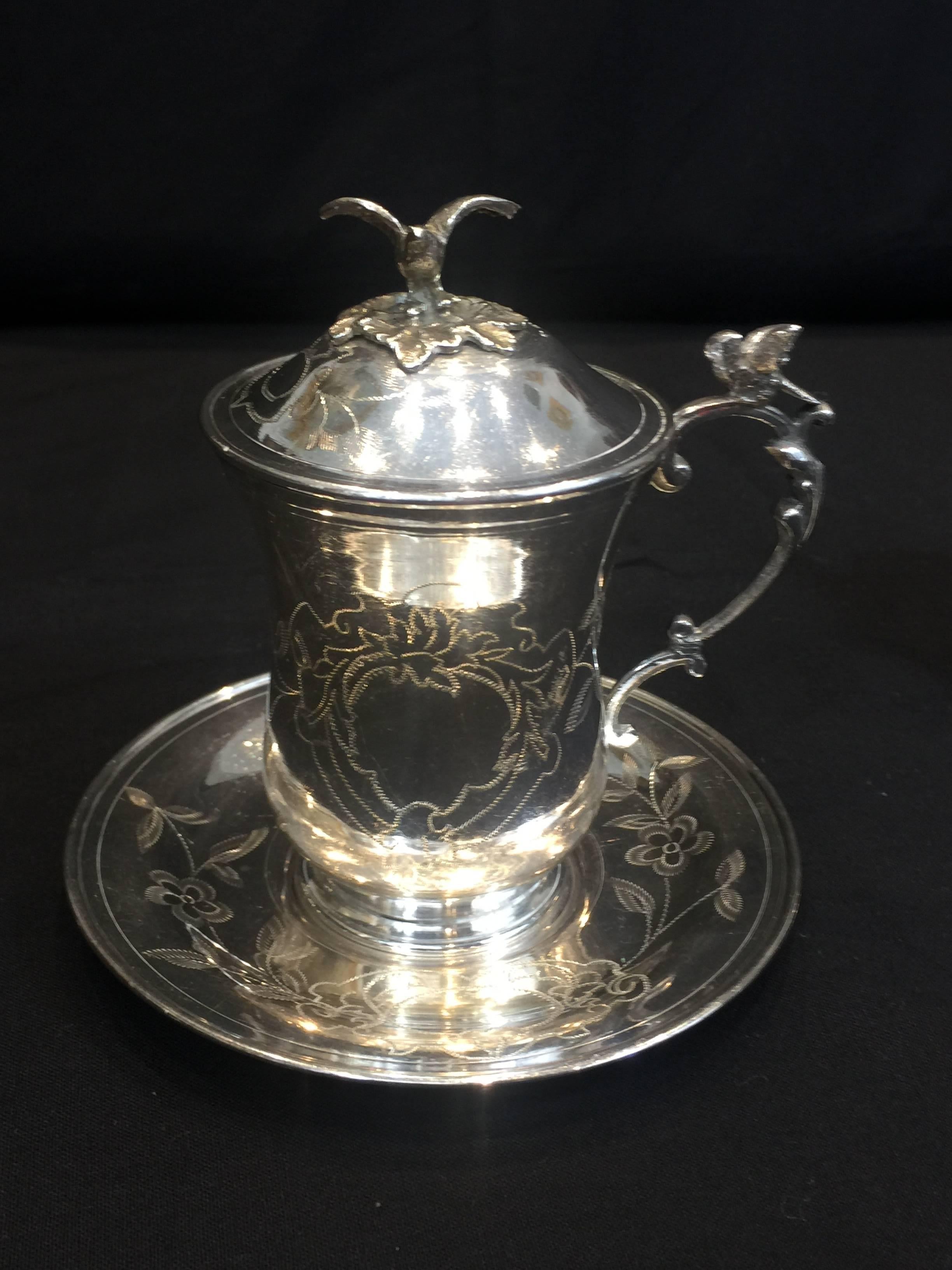A pair of Ottoman Empire style engraved silver sahlep cups. The cups feature decorative foliate shaped handles with birds perched atop, with the same birds with outstretched wings and leaves topping the lid. The lids, cups, and saucers are all