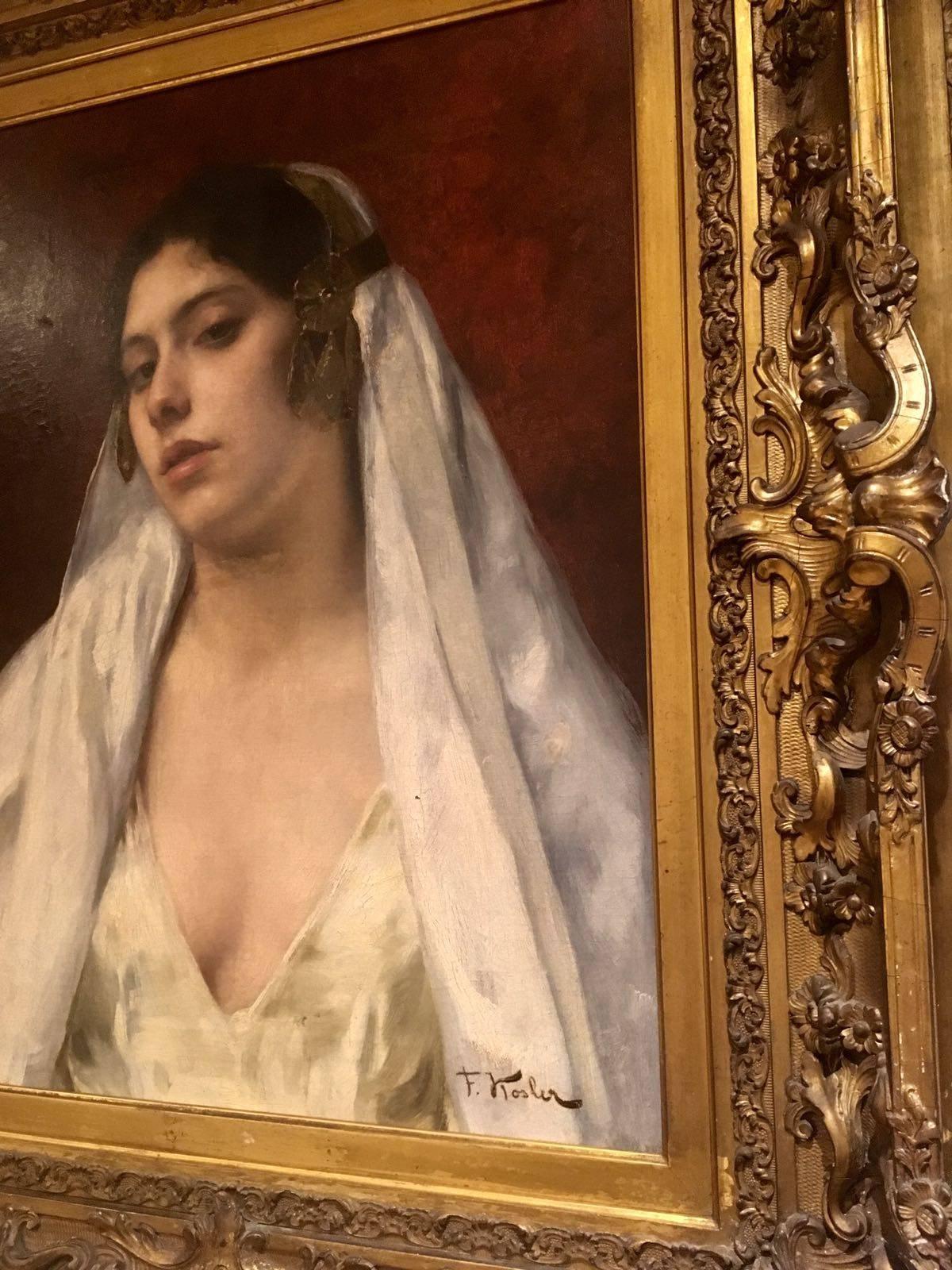 A striking portrait set on a dark reddish background of a young dark haired beauty wearing a cream colored low cut dress and a matching head scarf with side jewellery on the low headband holding it in place. She looks down and out at the viewer from