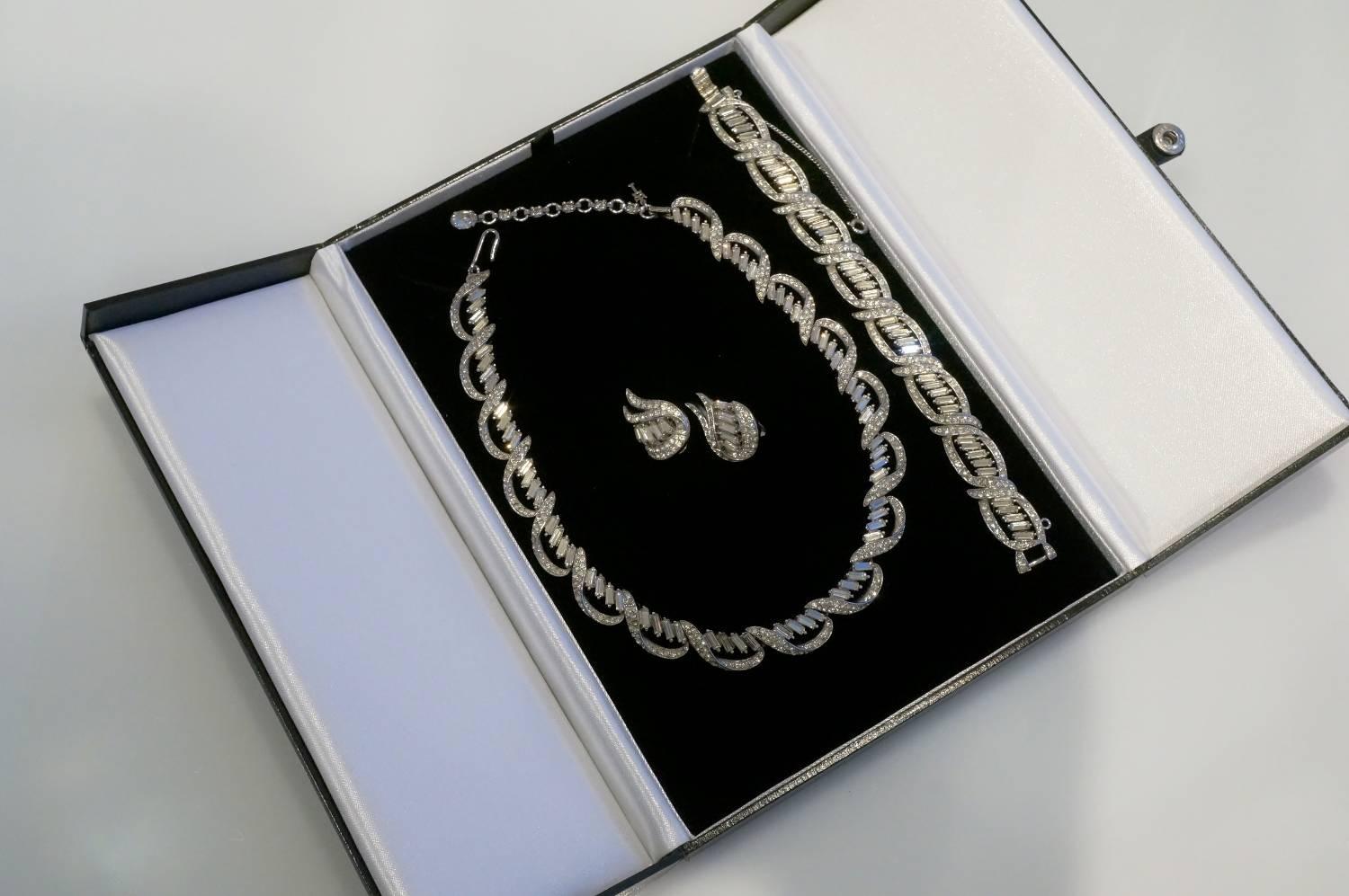 Trifari jewelry set necklace, bracelet and earrings, circa 1950s, USA. Baguette cut and chaton cut rhinestones on a rhodium plate frame. The necklace is finished with a large rhinestone and a Trifari key. The bracelet has a safety chain which is in