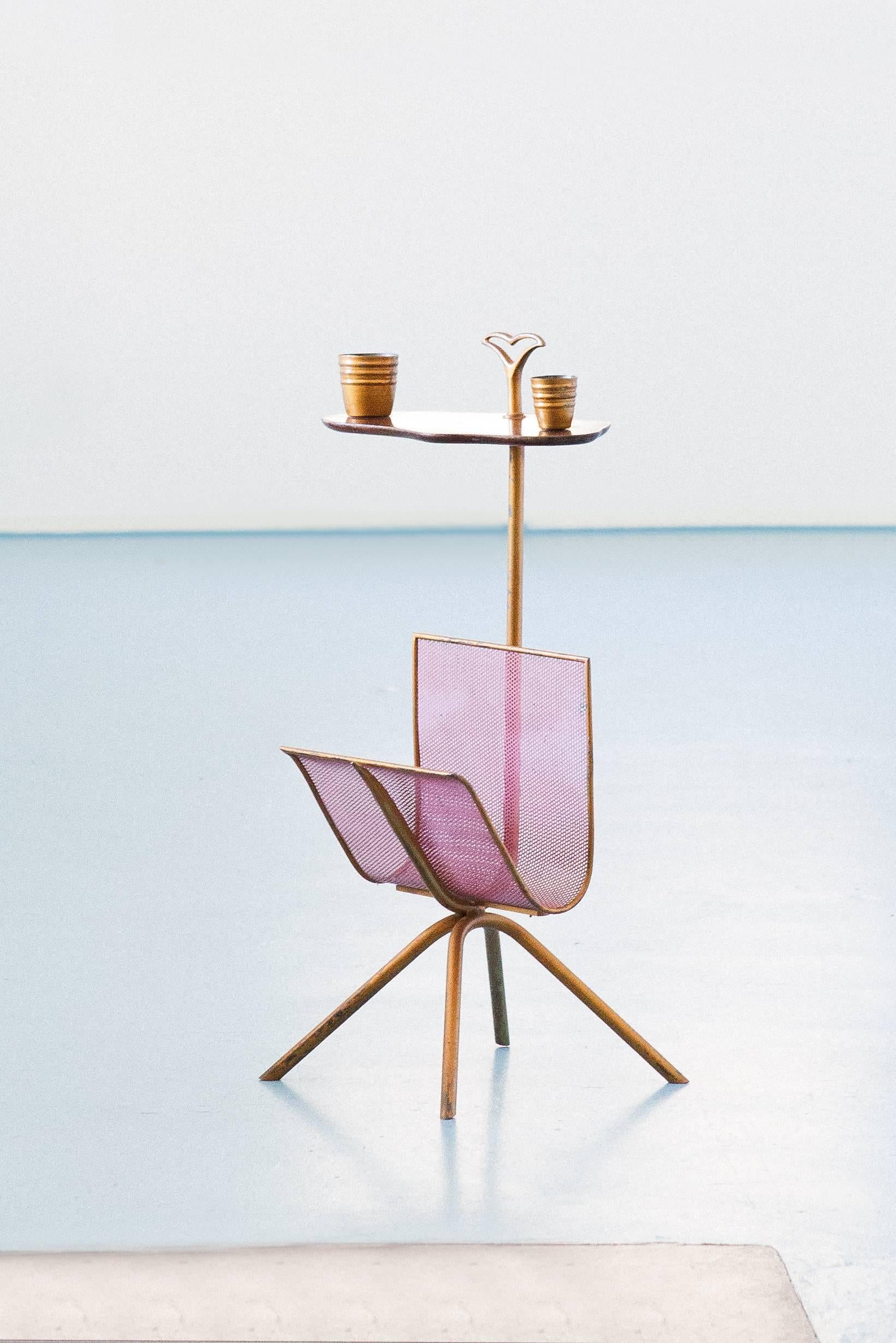 A side table with magazine rack , manufactured in Italy in 1950s
Solid brass gold paint finished frame with original patina , little mahogany wooden plane and four little brass glassess coordinated . 
The magazine holder is pink enameled .

This is