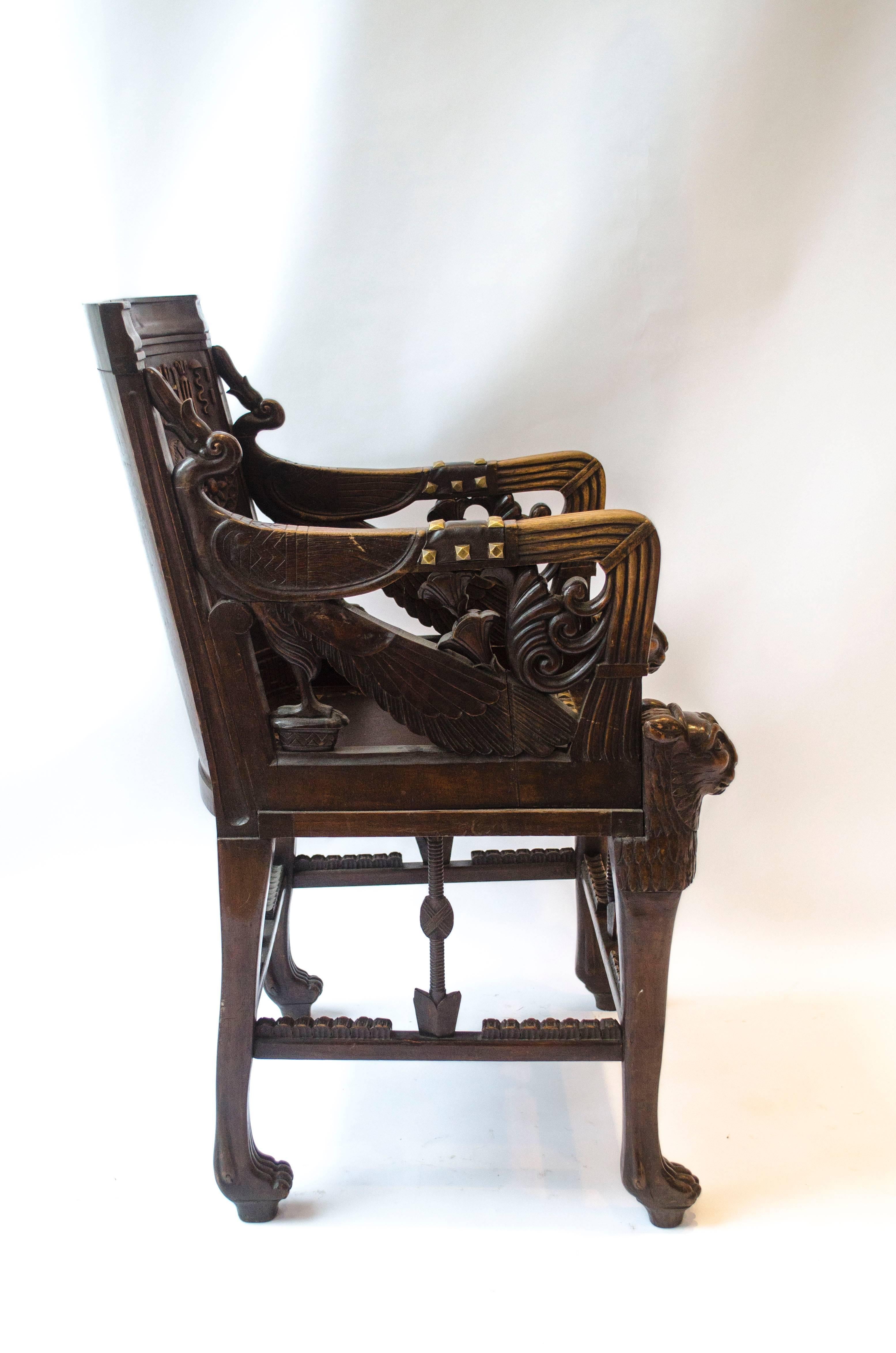 An early Egyptian Revival carved walnut elbow chair, the curved back with a pharonic figural scene, a leather seat and arm pads with brass studs, the front legs lion headed, on paw feet. Modelled on Tutankhamun's golden throne discovered by Howard
