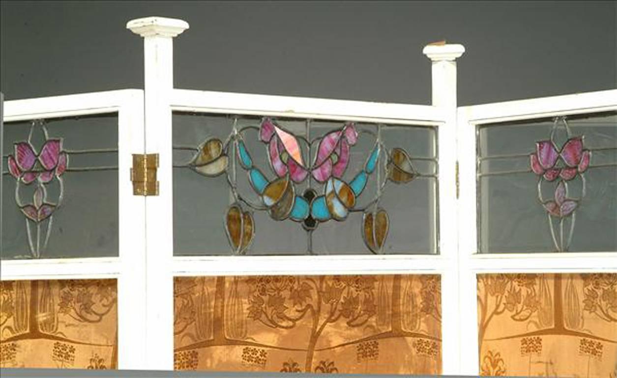 Liberty and Co. (attributed), a white painted enamel and stained glass three-fold screen, with capped uprights and stylised floral stained glass to the upper panels, the worn silk to the back probably designed by Arthur Silver of the Silver Studios