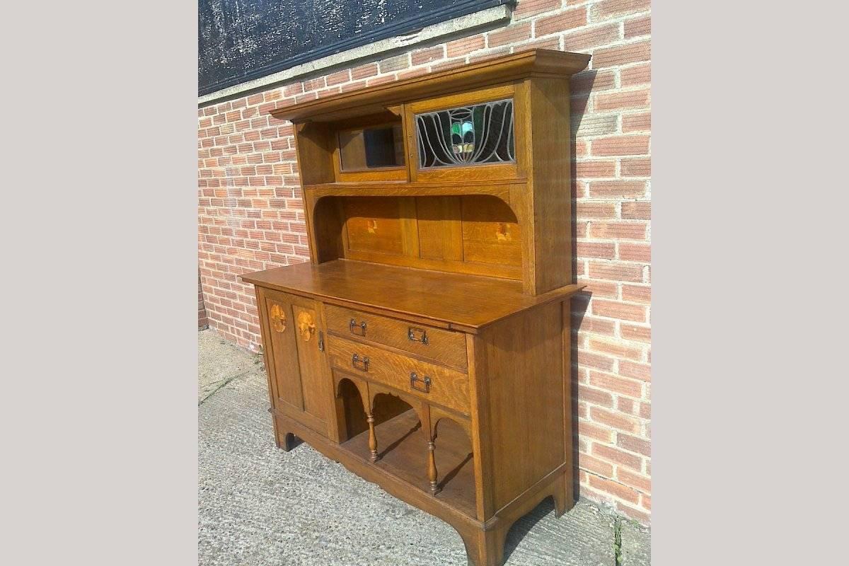 An Arts and Crafts Glasgow style Oak sideboard attributed to G M Ellwood made by Bath Cabinet Makers, with stained glass panels to the upper right hand cupboard, and stylised floral inlaid details with tulip wood, Stained Sycamore and Walnut above
