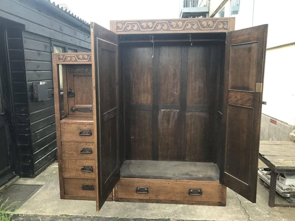 Liberty and Co.  attributed to Leonard F Wyburd,. A rare Arts and Crafts oak wardrobe with carved stylized floral details and hand wrought iron hinges and handles and a Cotswold style latch to the doors. A small cupboard to the left with four deep