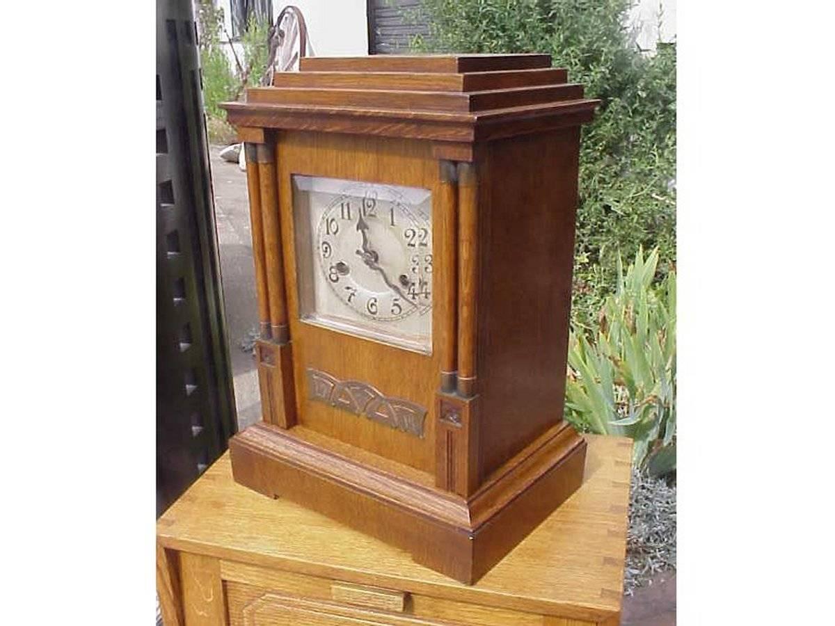A Continental Secession style Arts & Crafts oak mantle clock stepped top a silvered dial behind a full length door with bevelled glass flanked by two pairs of twin pillars with stylised brass mounts to the tops and bottoms with carved roses, and a