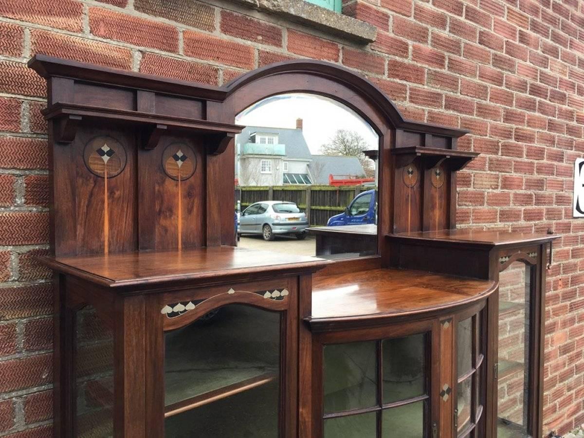 Une remarquable vitrine Arts & Crafts, avec des incrustations stylisées en ébène et en étain. Miroir biseauté en arc de cercle à l'arrière au-dessus de la zone de présentation, flanqué de deux étagères supérieures avec des détails marquetés stylisés