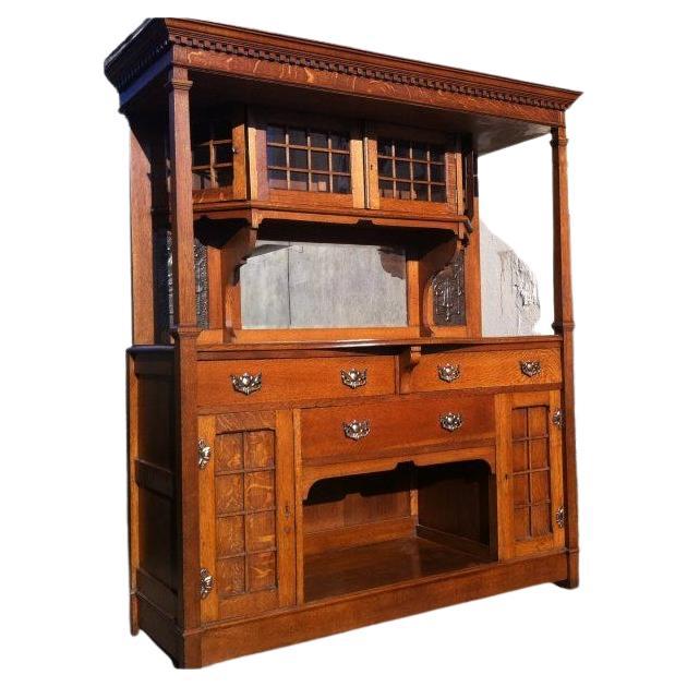 Liberty and Co attributed. An Arts & Crafts oak sideboard in the Glasgow style with a generous flat display area to the very top, dentil moulded cornice below with central upper angled cupboard and glazed doors, with beveled mirror below flanked by