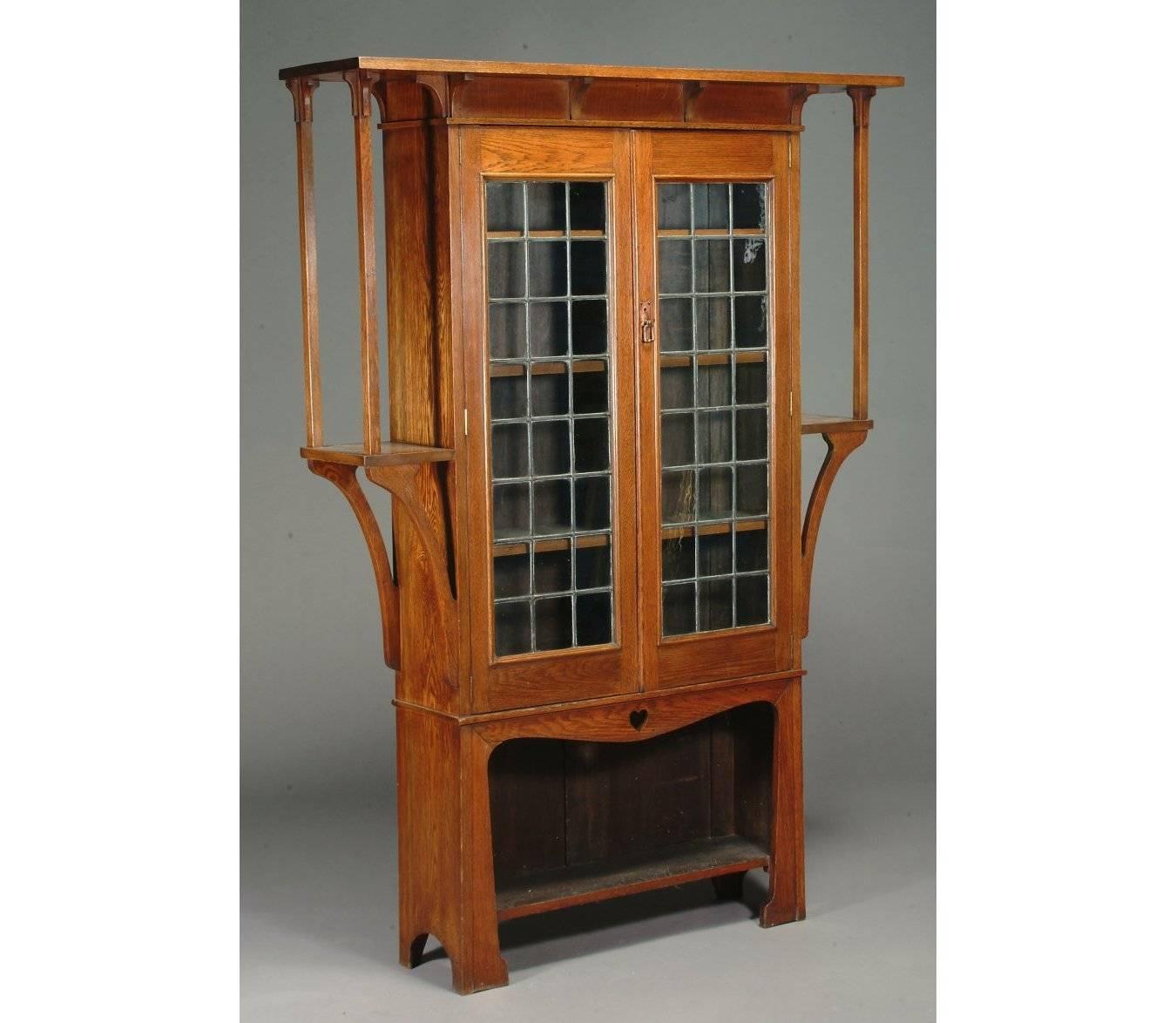 A near pair of oak Arts & Crafts bookcases by Liberty and Co. with two leaded glazed doors with adjustable shelves behind, with a single heart cut-out just above the lower open shelf. Extended shelves to each side with elongated supports uniting the