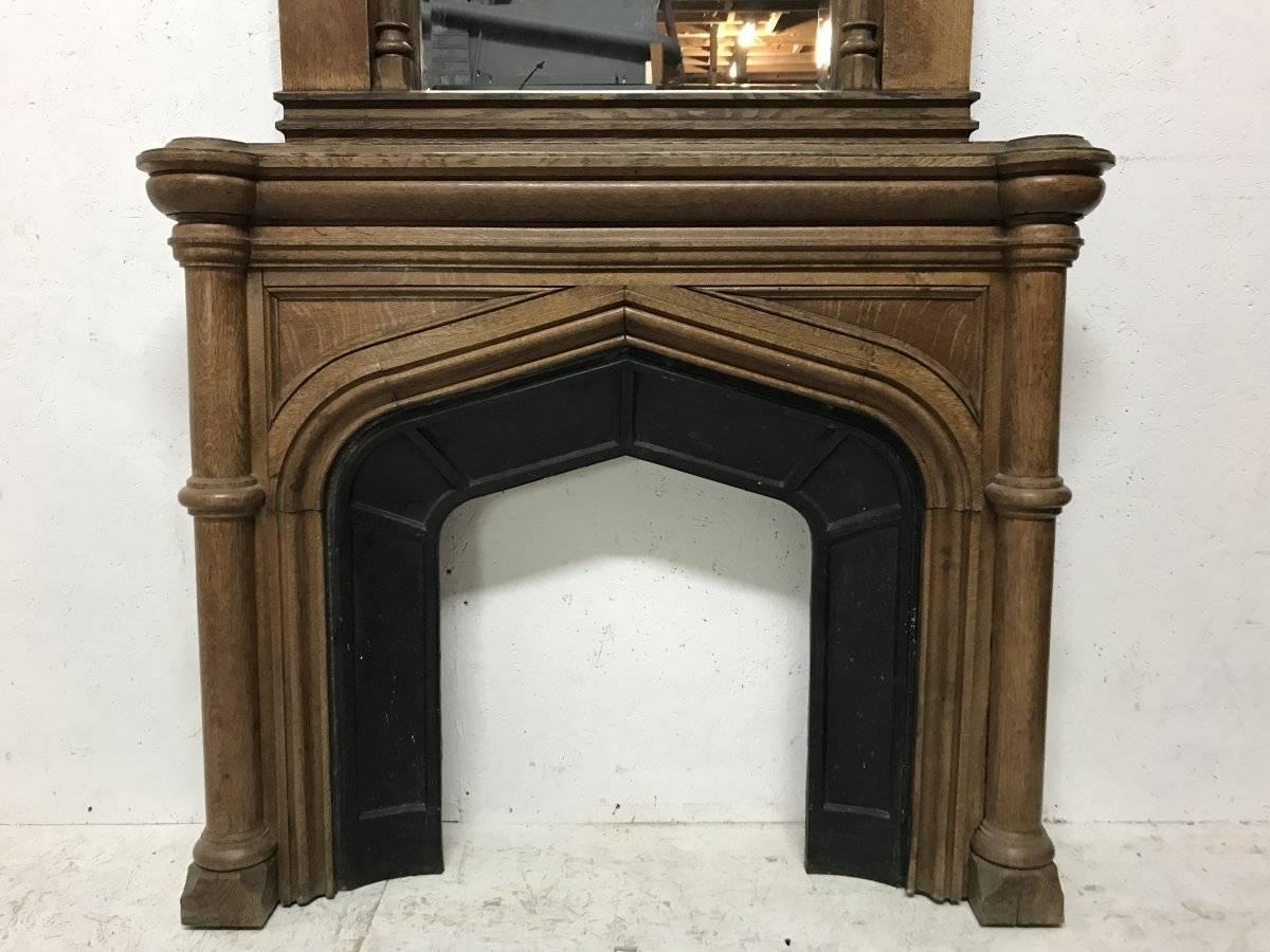 A complete French Gothic arched oak fireplace with its original overmantel mirror plate with projecting cornice with a wide oak frame and turned pillar details around the mirror and conforming larger turned details to the lower fronts. The sides