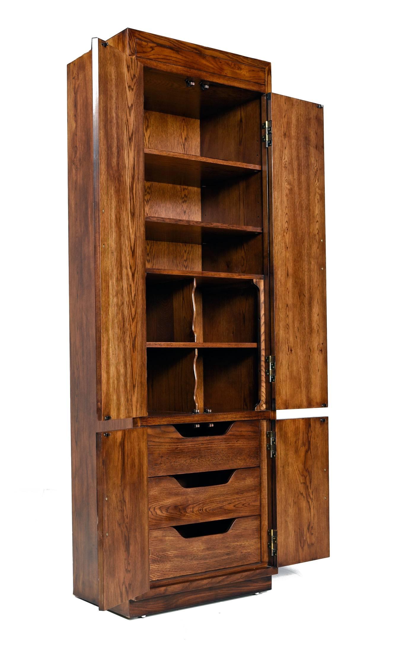 Vintage 1970s chifferobes by esteemed American manufacturer, Henredon. Handsome Campaign style pecan cabinets are accented with brass hardware. 
Open the cabinets to reveal a combination of drawers, cubbies and shelves. Both open shelves and