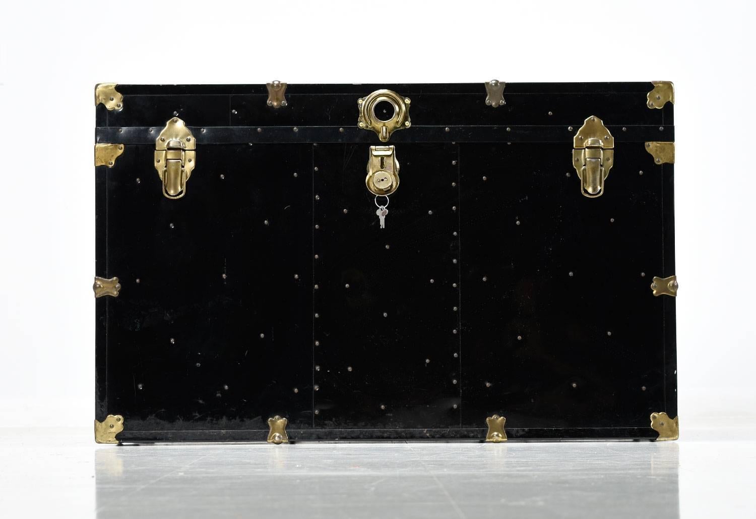 Vintage black enamel over metal steamer trunk accented with leather strap handles and gold colored latches. Open the trunk to reveal removal storage trays at top. The interior is a radiant blue. The trunk comes with a key.