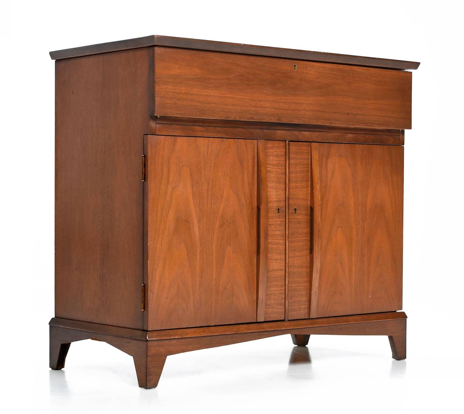 Midcentury American made walnut bar cabinet. This unassuming piece looks like a typical server when closed. Lift the top to reveal a Formica top surface for preparing drinks, and several divided spaces for storing glassware. The lower portion