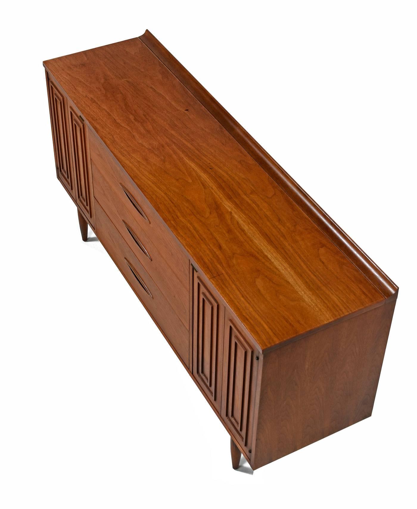 Mid-Century Modern sculptra credenza by Broyhill Premier. Back edge of top surface is accented with an upward reaching sculpted lip. Signature relief cut pattern adorns the cabinet doors. Inset elliptical handles on the drawers enhance the atomic