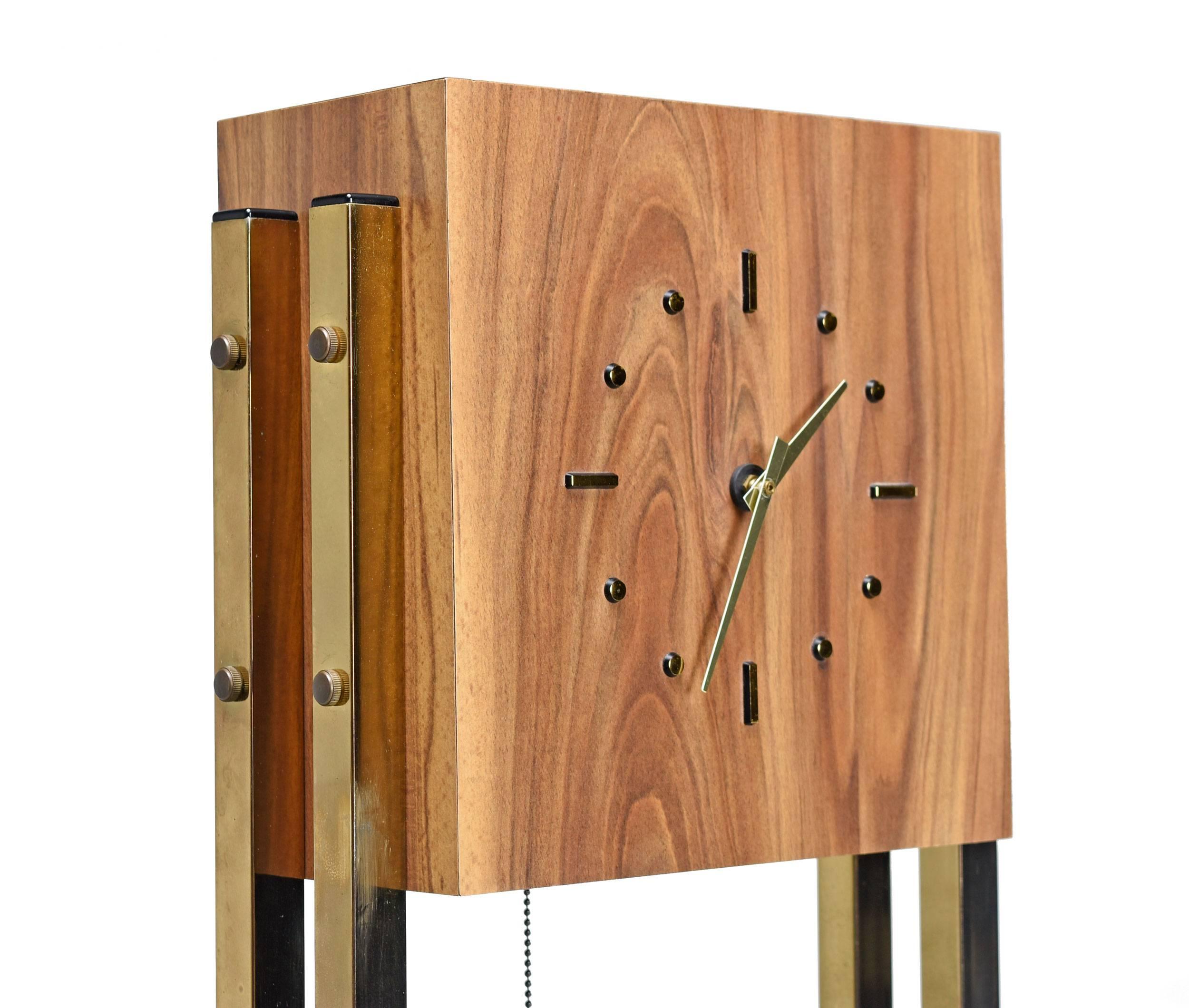Vintage 1970s grandfather clock with Minimalist modern design. The sleek and sexy clock features brass pillars and a laminate surface that mimics the look of wood. A light emanate's from beneath the clock face with a convenient chain switch. The