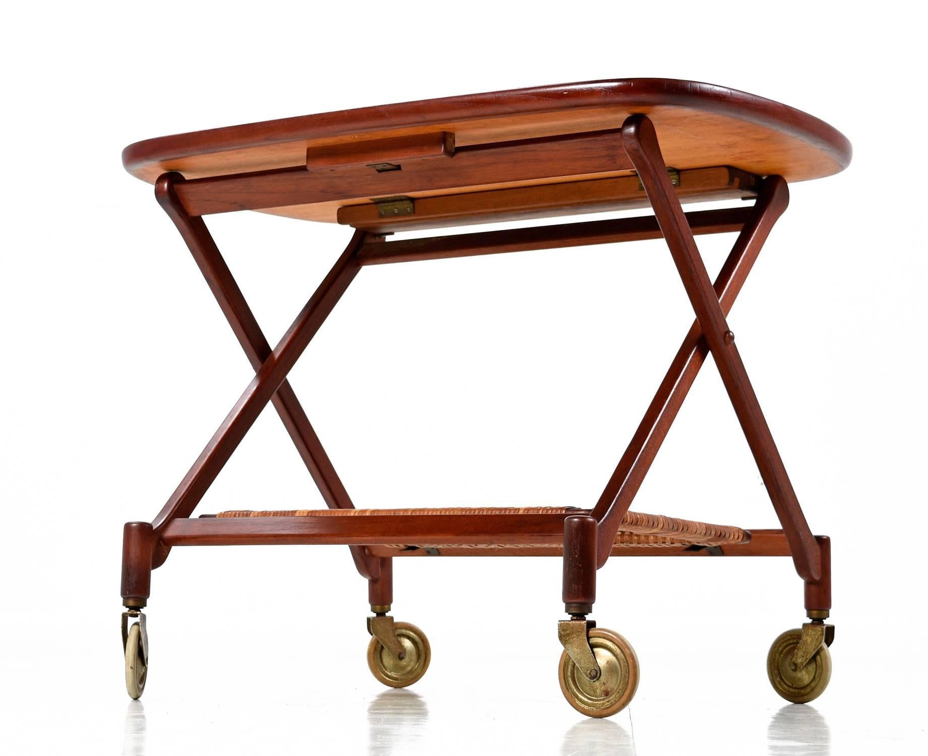 Poul Hundevad for Vamdrup folding teak bar cart. This early Scandinavian gem is a rare piece by noted designer Poul Hundevad. The lower level features a woven cane shelf. The oval tray has an elevated lip surrounding the perimeter. The cart is
