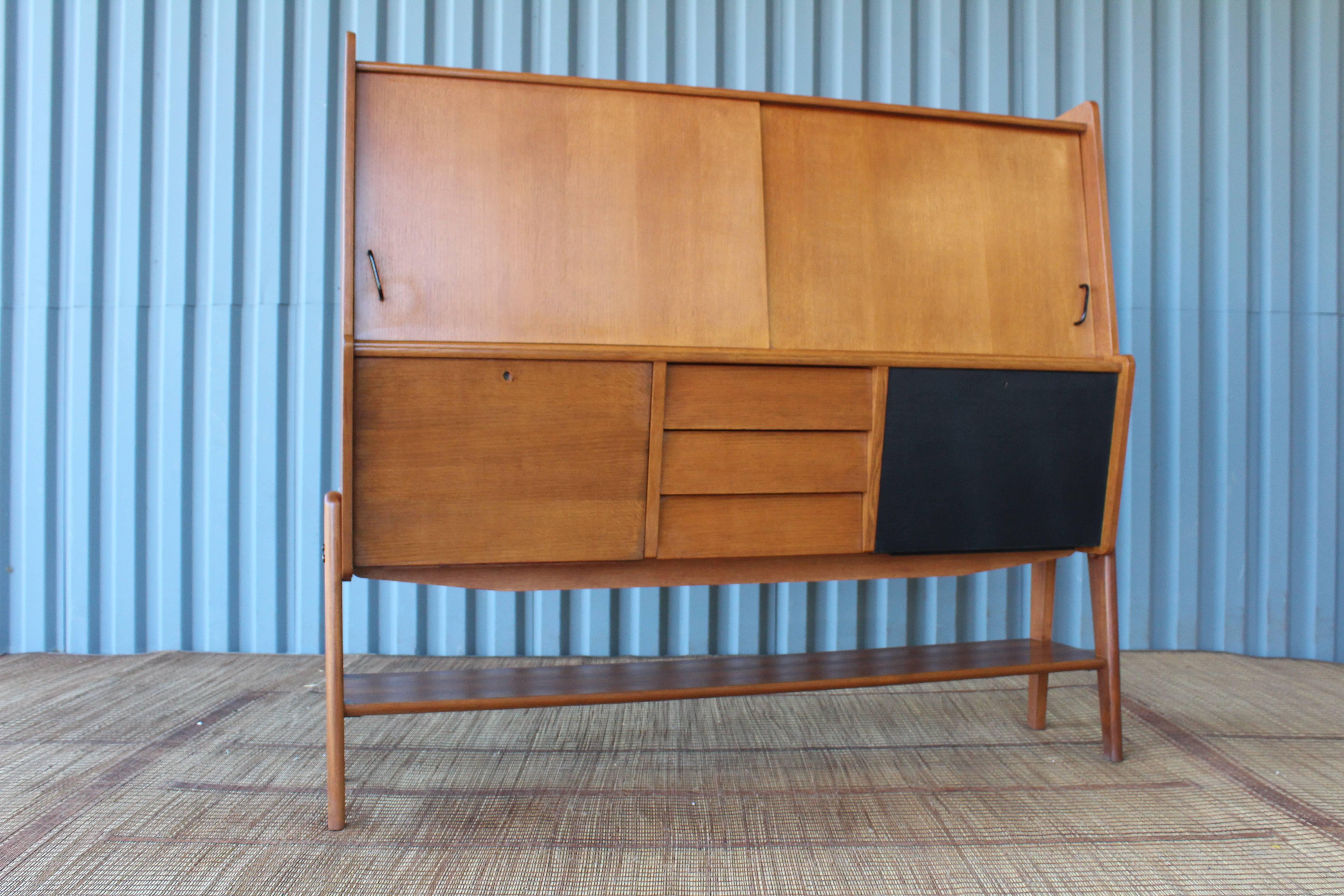 A 1950s teak high board credenza. This piece offers loads of additional storage. Features sliding doors, three dovetail joined drawers, and two drop down compartments- one of which can function as bar to hold tall bottles and shelf for cocktail