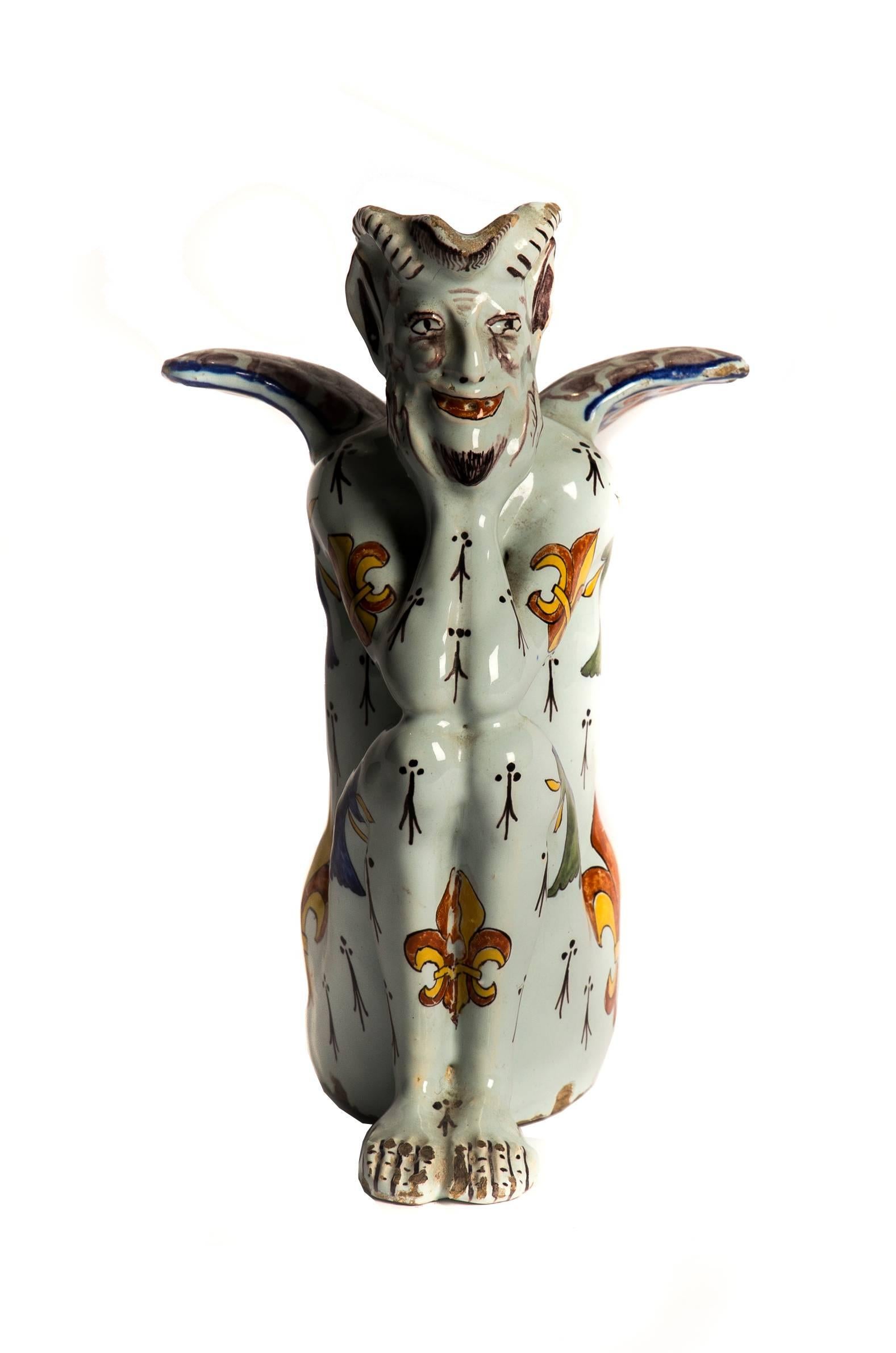 A rare tin glazed pottery devil with exquisite decorative details including fleur-de-lys and oak leaf motifs and unusual repeated black symbols. 

In enigmatic seated pose with elbows on knees and characterful grin. Beautiful detailing on the wing