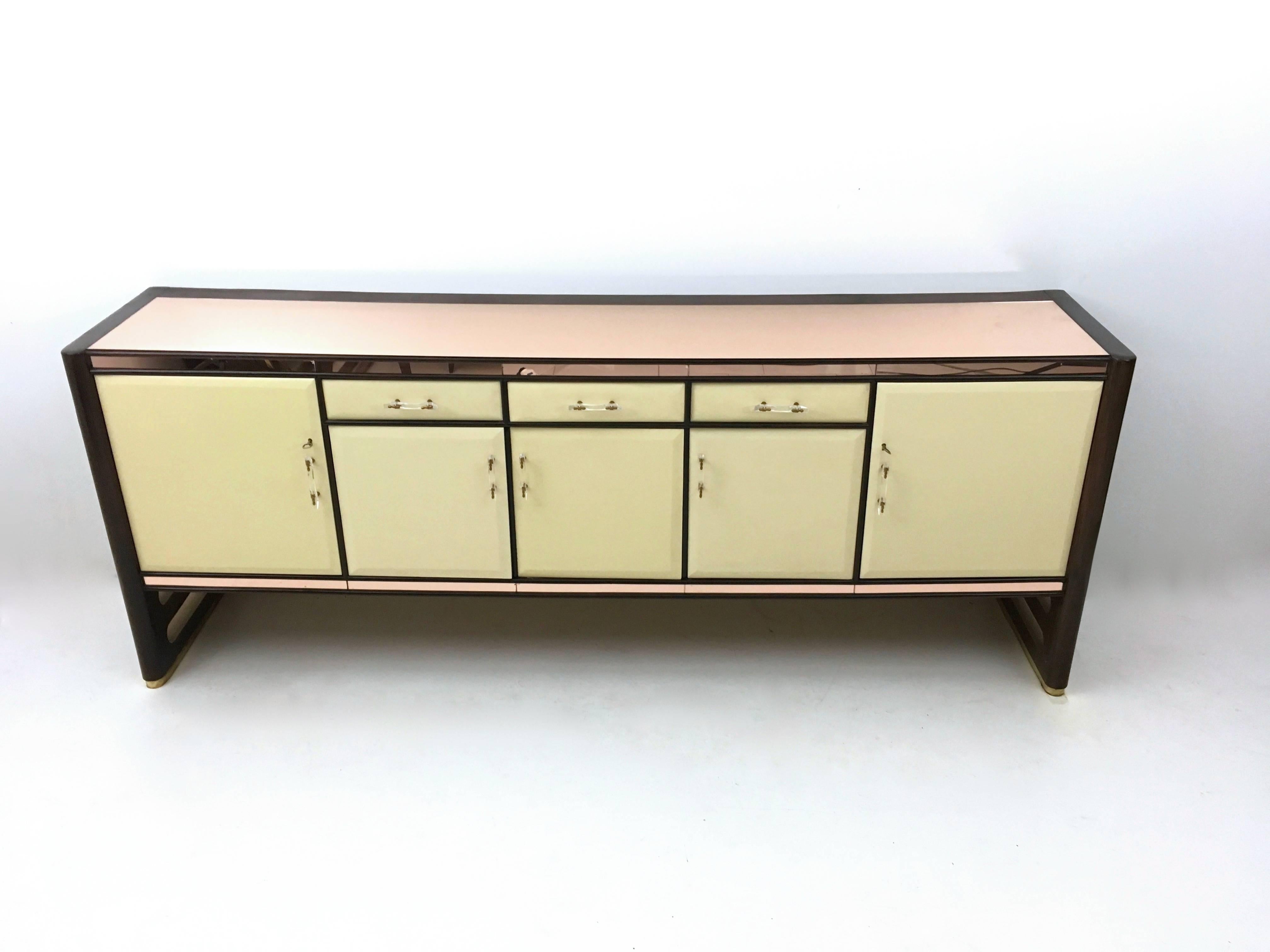 This sideboard is made from wood, with lacquered wood doors, a pink mirror top, brass and perspex handles and maple interiors.