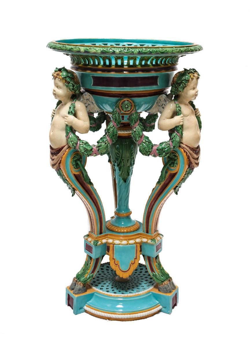 A Minton England majolica jardiniere featuring three faun footed cherubian figures serving as pillars supporting a reticulated planter top. Each winged cherub connected with a ribboned vine leaf draped around their neck.

Impressed marks for