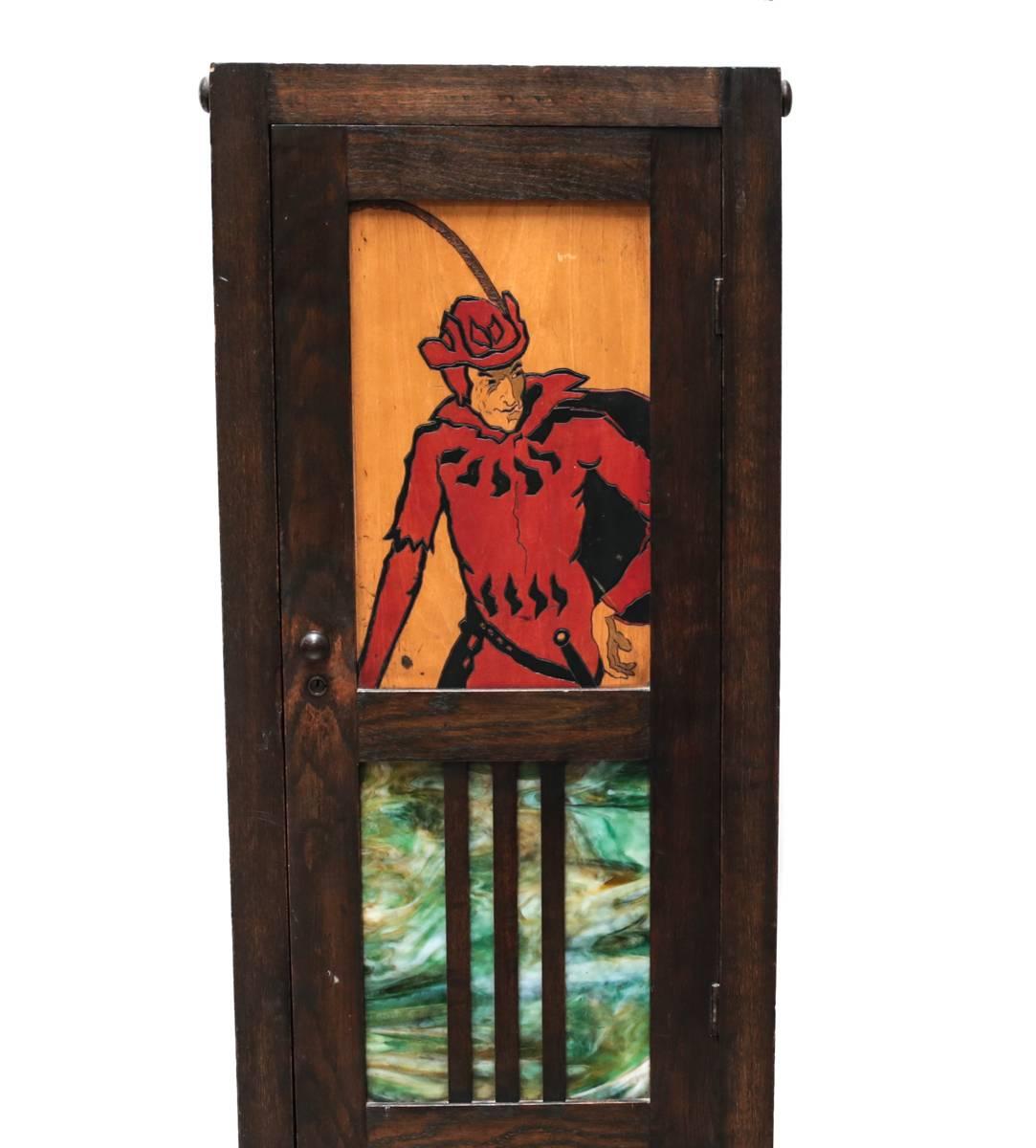 An unusual early 20th century stained oak cabinet, the front door set with a colorful slag glass panel and with a painted and engraved figural depiction of a musketeer or Robin Hood type on an inset panel of maple wood. 

Opens to reveal a three