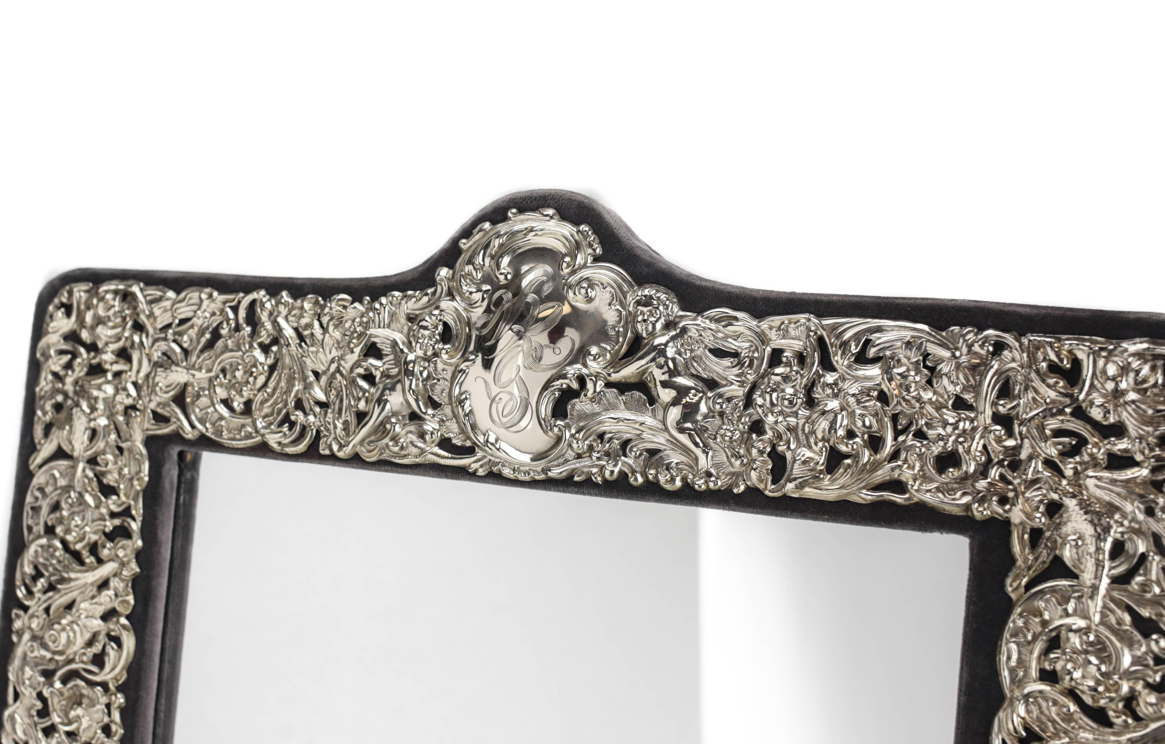 A large American table mirror with an ornate sterling silver mounted border depicting a vine and floral arrangement with two winged cherub’s center top, set on a grey velvet back with easel back. Late 19th or early 20th century.

Lower right