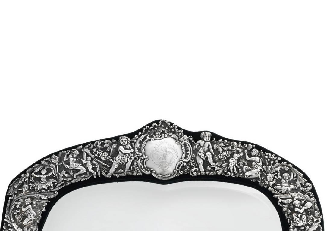 A large sterling silver cast and chased border decorated mirror frame mounted on a green velvet wrapped wood back with hinged Stand. Depicting foliage and grotesques, the top centered by a Rococo cartouche engraved with coat of arms and flanked by