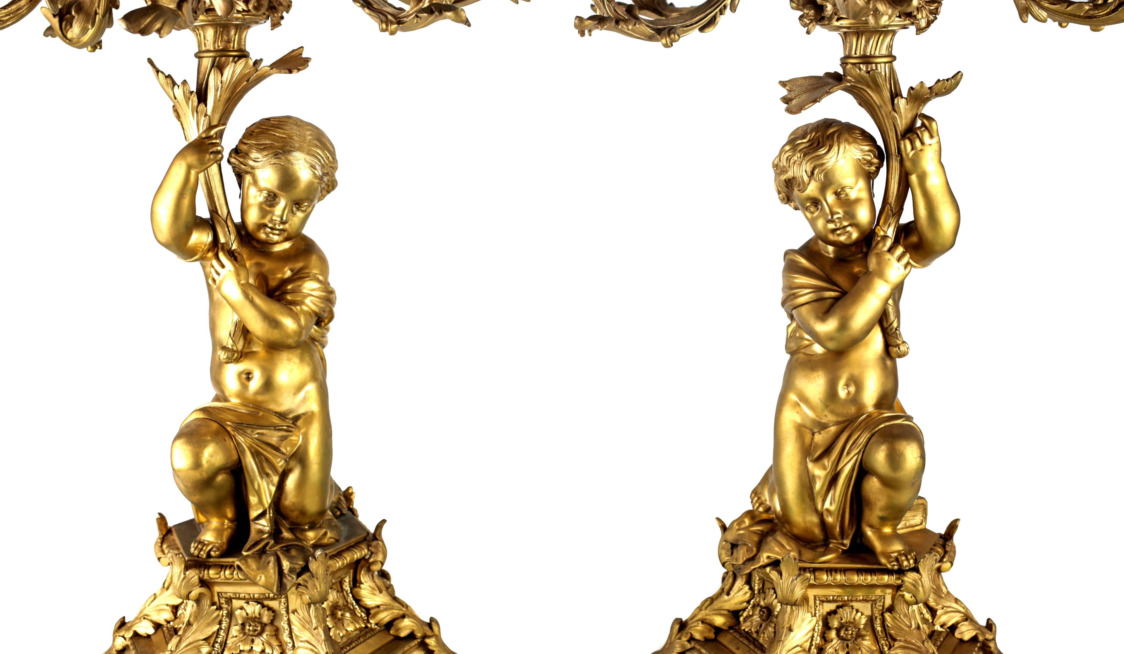 A fine and large pair of French gilt bronze candelabras each stationed with a prominent figure of a putti or cherub raising ten acanthus cast candle arms. The cloth draped figures sitting atop a stack of books, the hexagonal shaped base with foliate