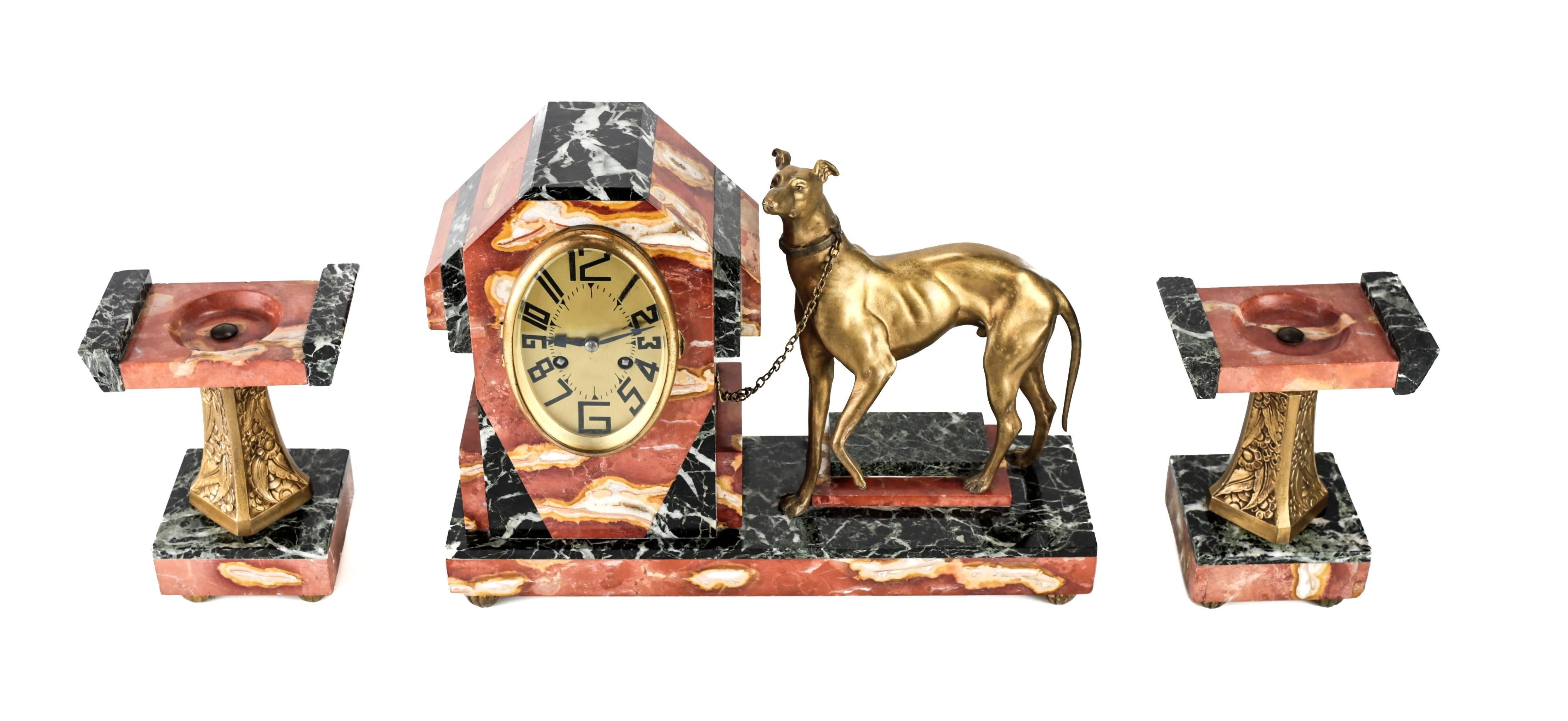 An Art Deco three-piece clock garniture with gorgeous red and black marble and gilt French bronze greyhound. The dog chained to the elongated oval clock housing. Matching garnitures, each with floral decorated columns. 

The centre piece: 16