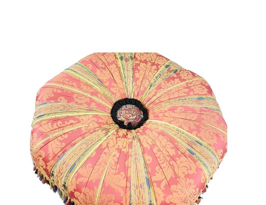 An eclectic and vibrant upholstered tuffet ottoman by Mackenzie Childs. Beautiful pink floral fabric with multicolored tassels. Stacked ceramic bun feet with various hand-painted pattern designs. Bearing the maker's metal tag on the underside.