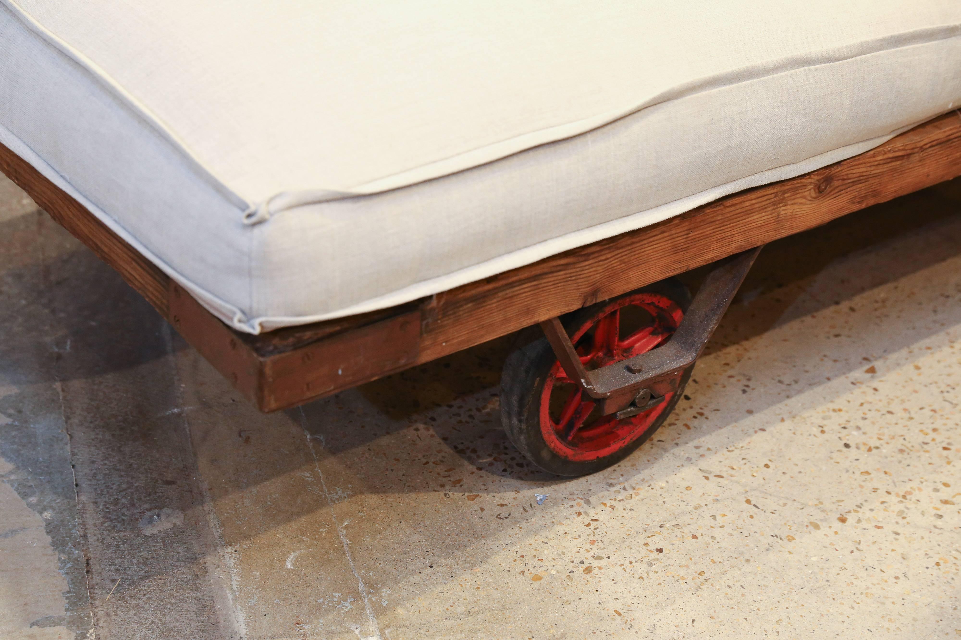 Piled with pillows this Industrial factory trolley becomes a chaise or a sofa but, when the pillows are removed, it instantly converts into a daybed. Three heavy duty wheels with original red paint support the heavy wooden frame with iron corner