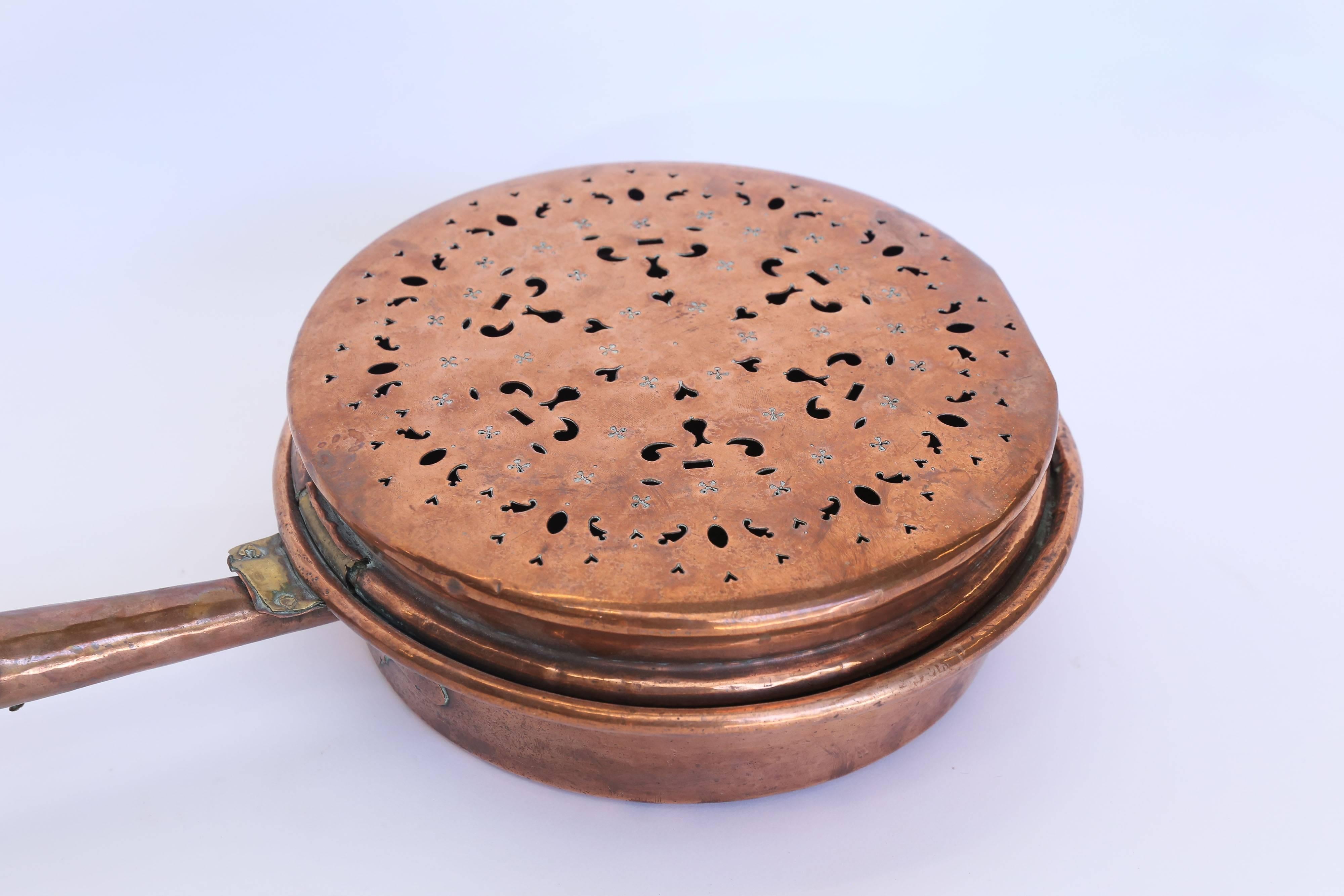 From France, a chestnut fireplace roaster made of copper with a wooden handle. The hinged lid is reticulated with oval, heart, and comma shaped piercings. The pan has some dings and dents and is in typical condition for an item of its age.