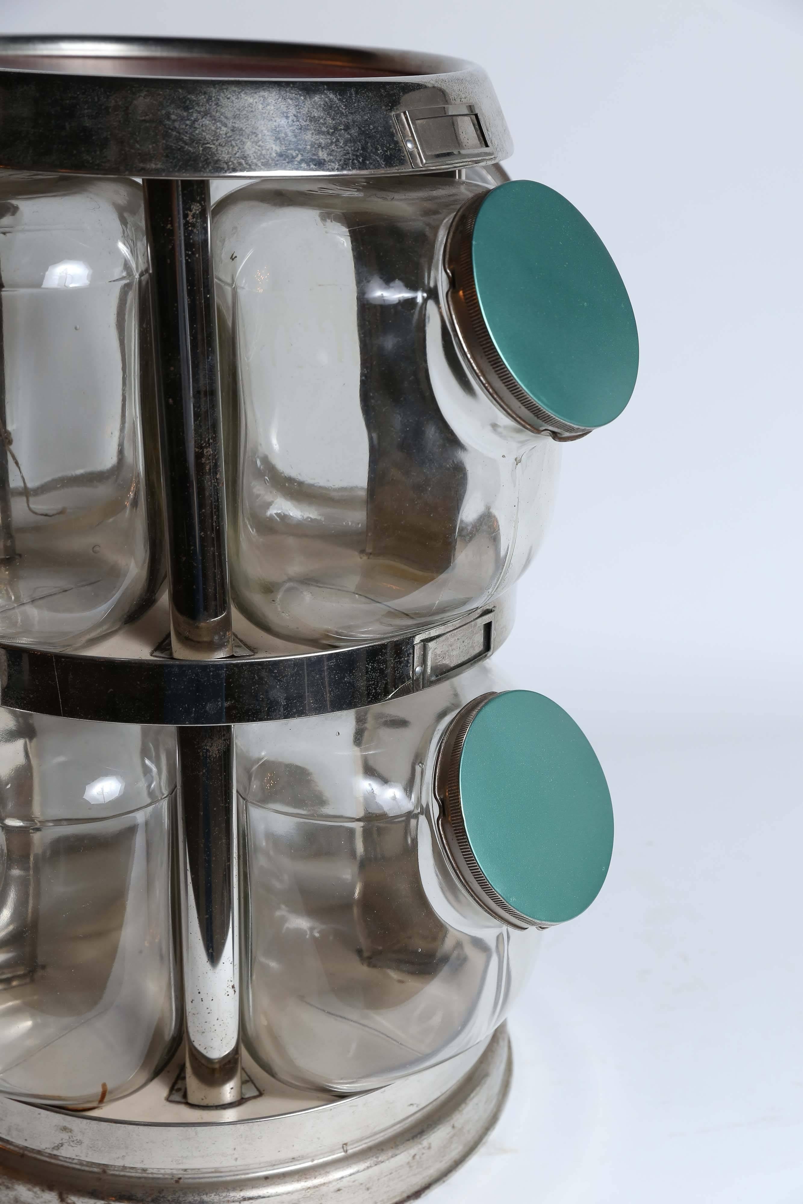 Eight glass jars with green metal lids and a red top surface in a chrome revolving Stand. There is a metal slot above each jar for identifying information. The metal has slight corrosion and could use a good cleaning but that is best left to the