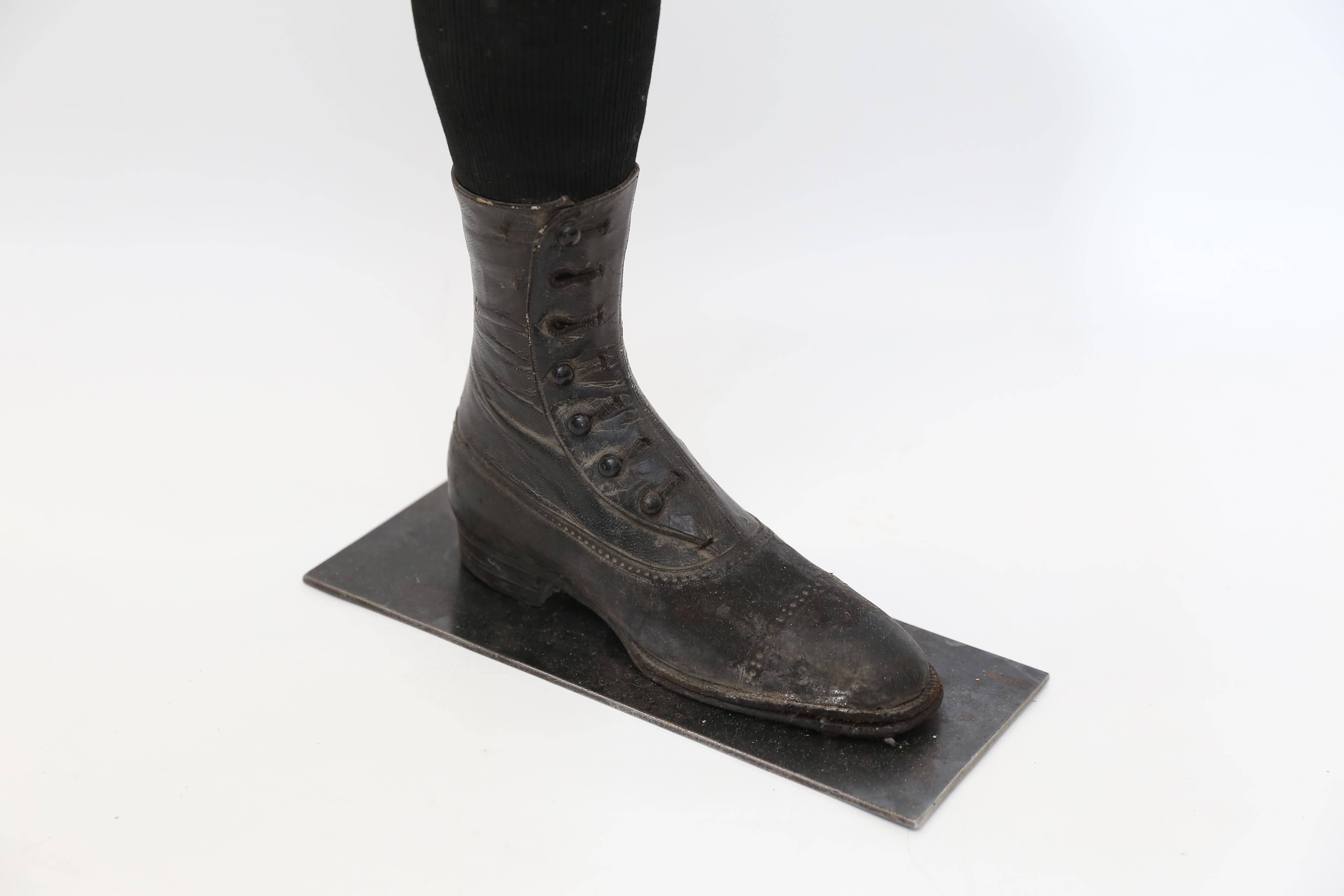 The leg of a wooden mannequin with a child's high-top shoe and black ribbed sock from the Victorian Era mounted on a metal base. The seven button shoe is of finely stitched leather with a hard leather sole. Two of the buttons are missing. Mounted on