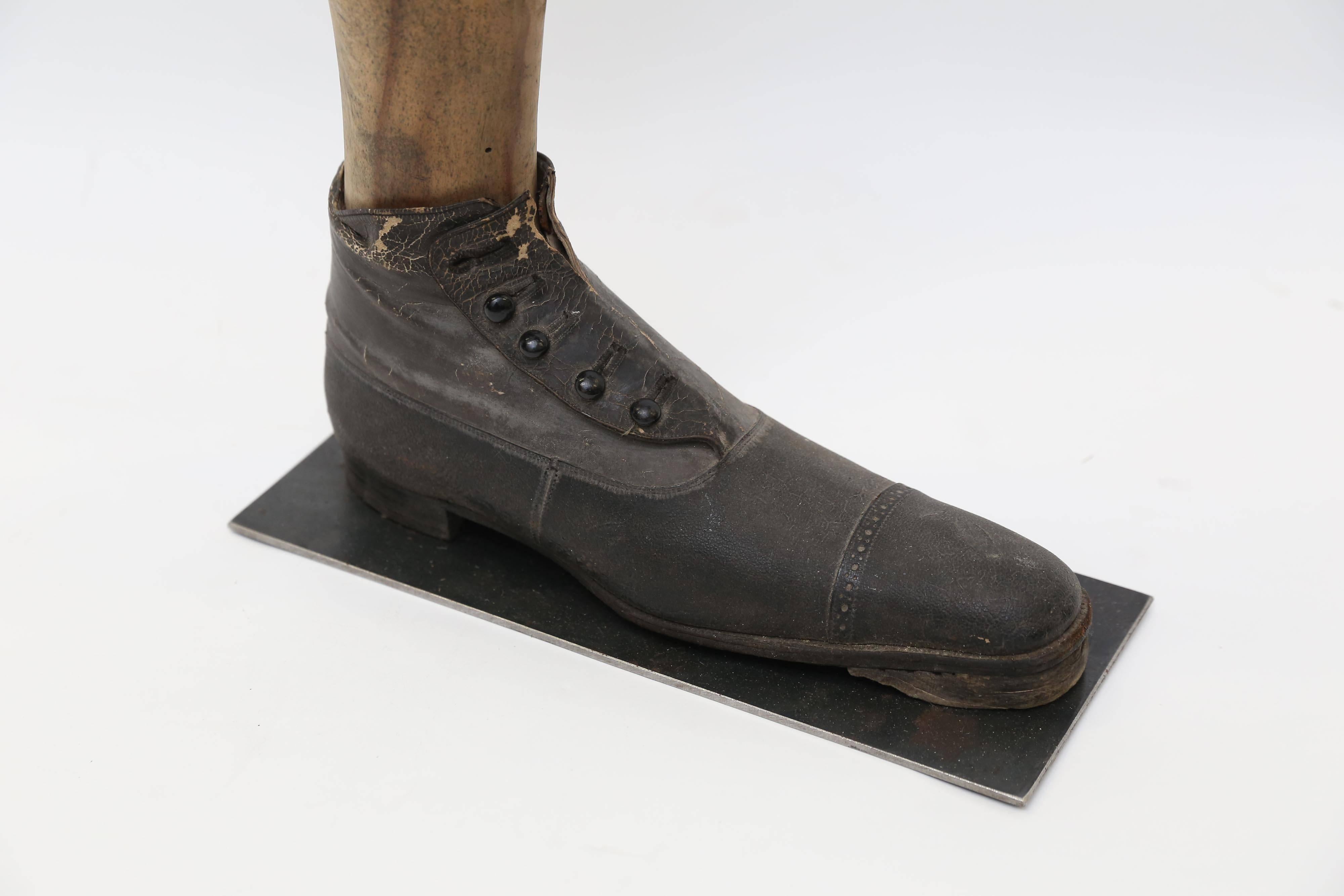 The leg of a wooden mannequin with a man's button-up shoe from the Victorian Era mounted on a metal base. The five button shoe is of finely stitched black leather with a hard leather sole. One of the buttons is missing. Mounted on a custom steel