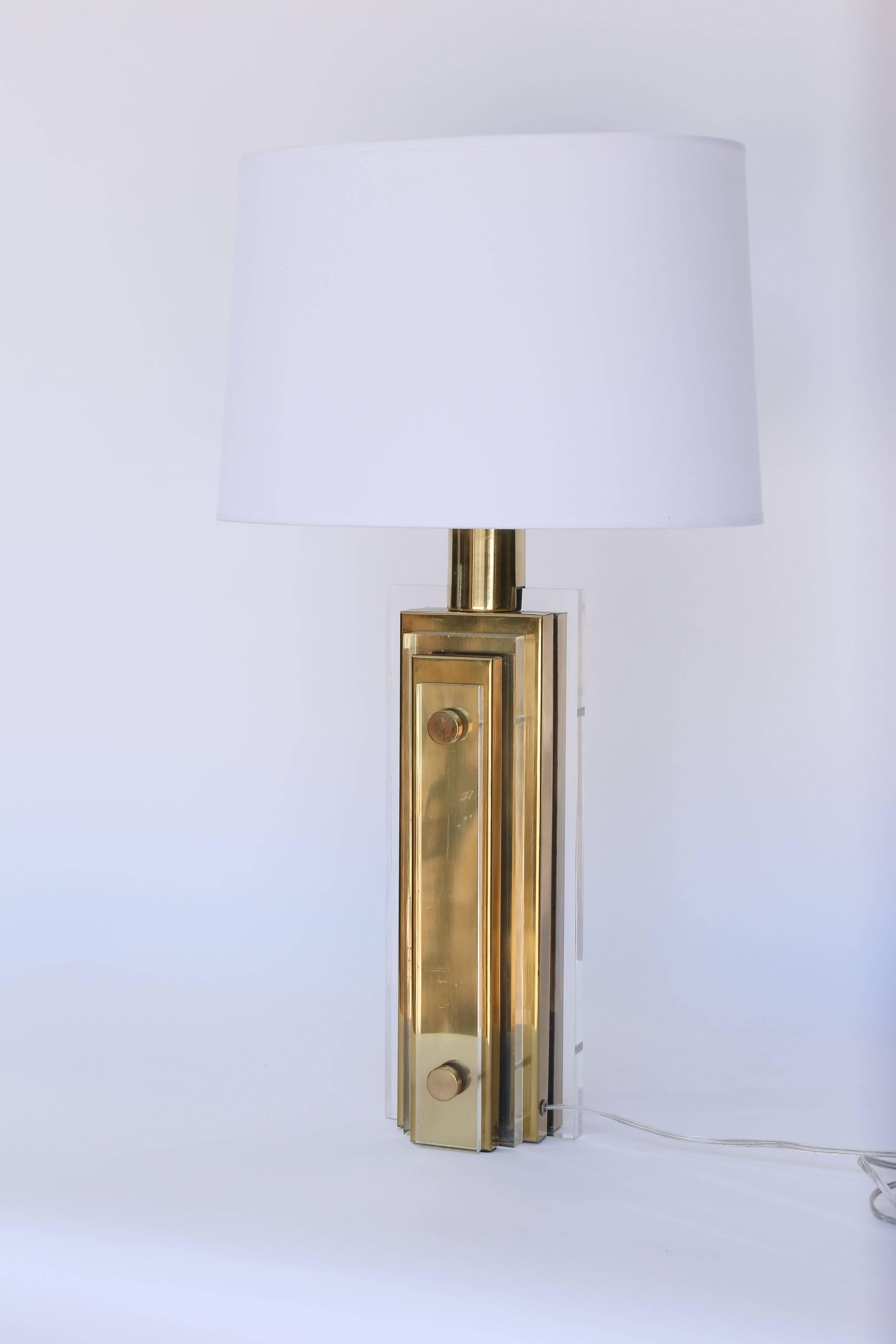 A dramatic brass and acrylic table lamp reminiscent of the Cityscape style of Paul Evans. Alternating slabs of brass and acrylic of ascending heights are pierced by two brass rods. Topped by a white oval shade measuring 9.5 inches by 15 inches.