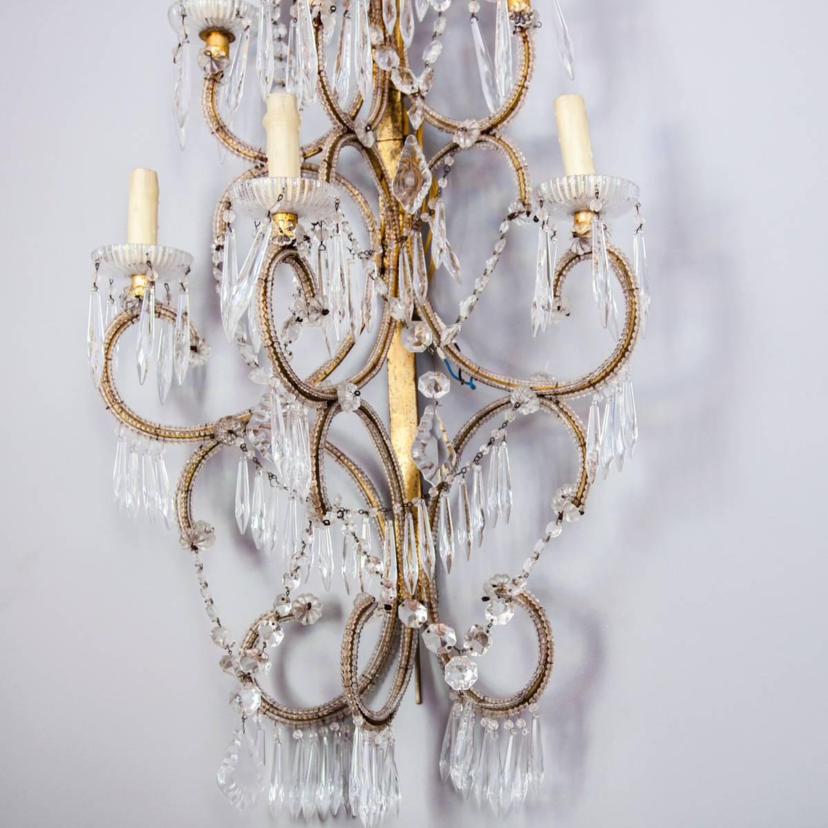 Pair of sconces, in the Italian Maria Theresa style, each having a frame of beaded gilded iron, six scrolling candle arms adorned with beading, terminating in scalloped glass bobeches, the entire fixture draped in swags of crystal strands, prisms
