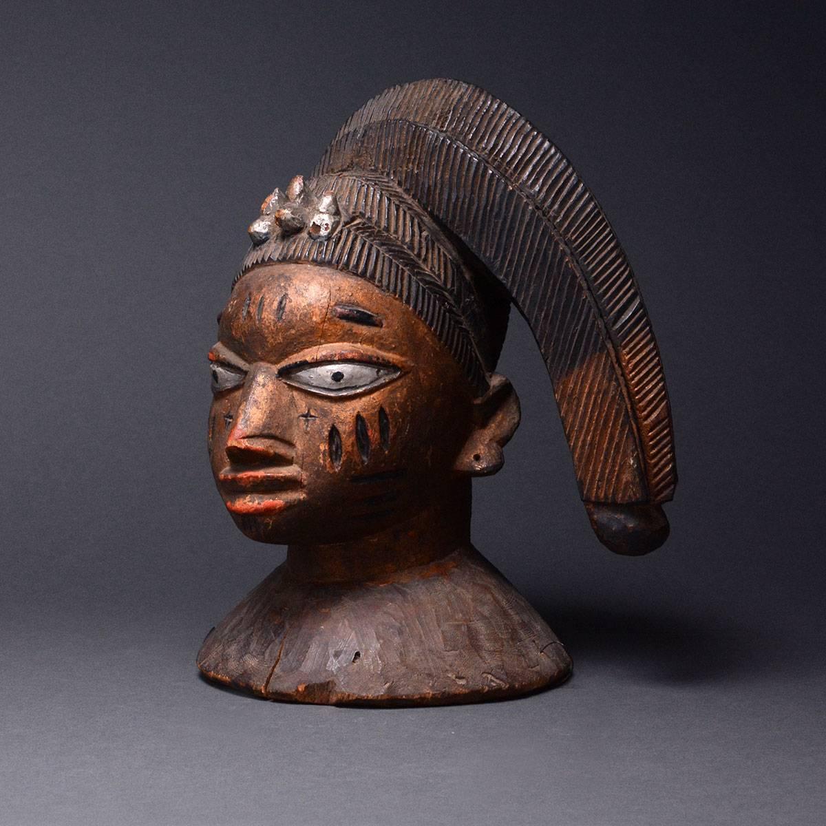 This attractive and ornate mask was made by the Yoruba. The base is a small, flat pyramid of wood, giving rise to a columnar neck that in turn supports the head. The face is rounded with a pointed chin, hooded eyes, a flat nose, a wide mouth and