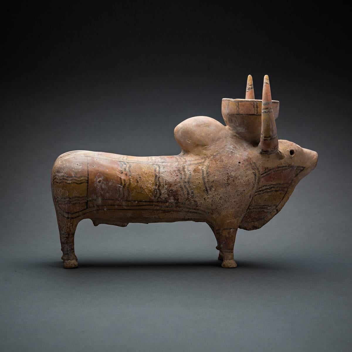 Large humped terracotta zebu bull standing on the four legs, his head supporting between two solid up-bent horns what looks like a ceremonial basket, the whole surface painted in bright colourful geometric patterns. The snout carefully sculpted with