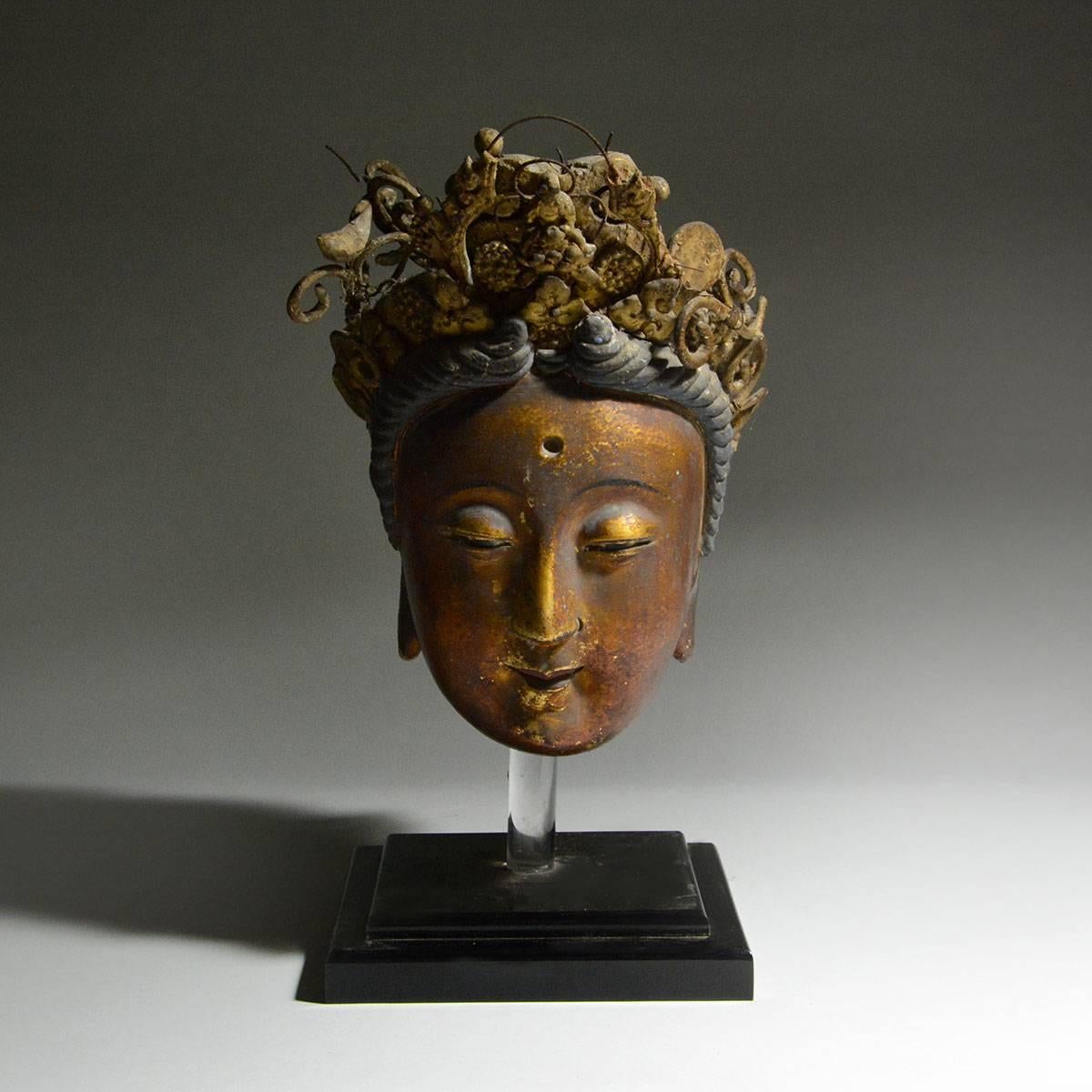 Cast in exquisite detail, this sculpture of Kuan-Yin is applied with red and black lacquer and gilt in the face and black and blue pigment in the headdress. Her oval face, aquiline nose, pursed lips, and delicately shaped downcast eyes make up her