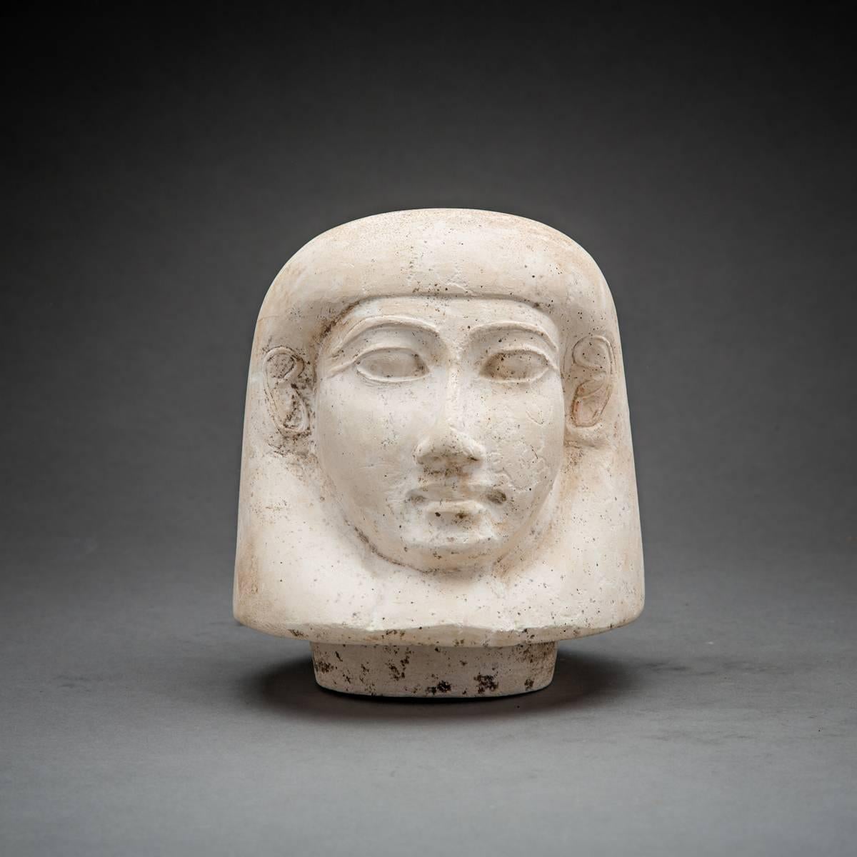 The treatment of this head as an independent unit with a circular, recessed projection on its under side suggests its use as a lid on a Canopic jar. The features of its cordiform-shaped face are idealing and are dominated by
