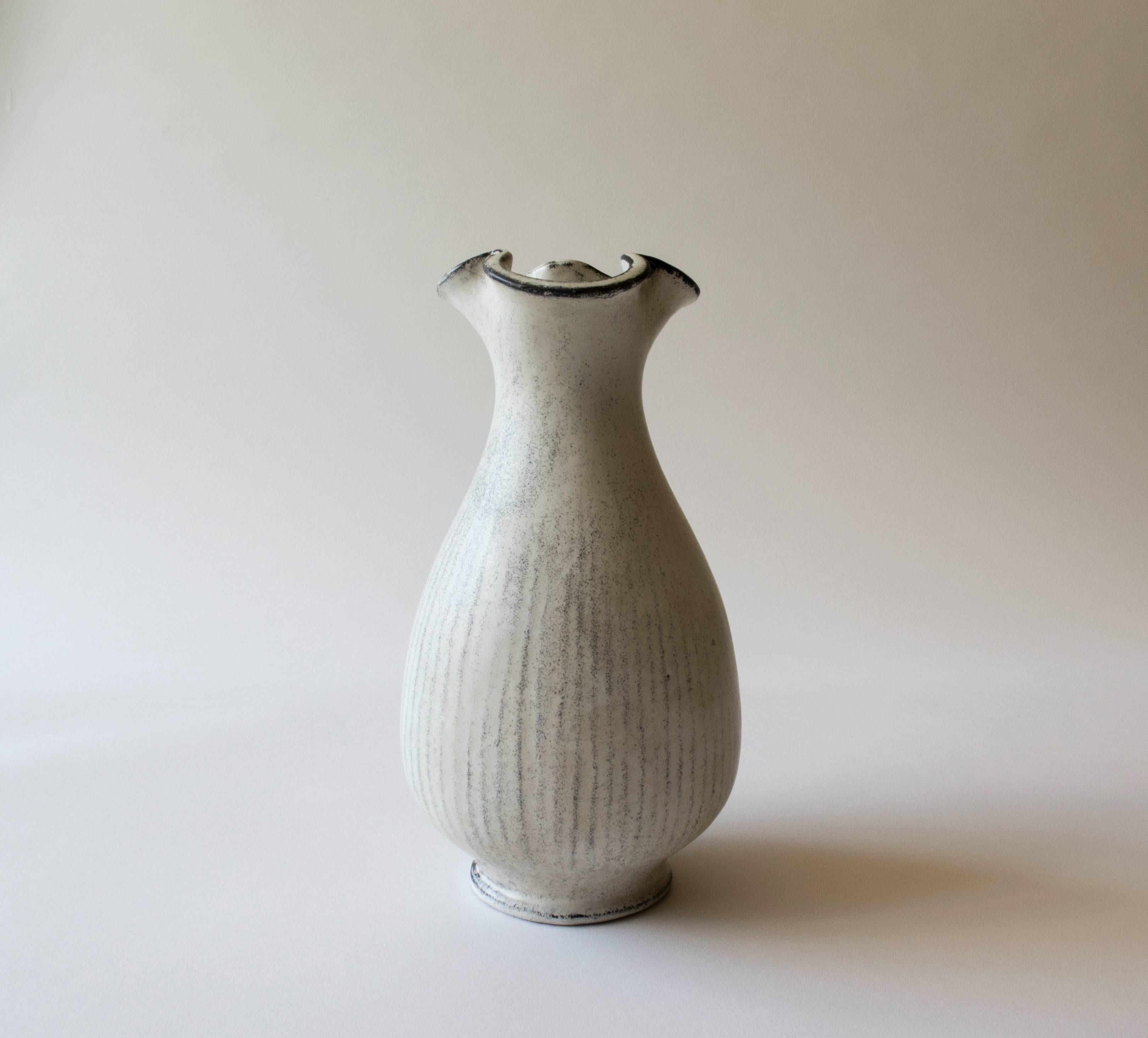 An elegant and refined form. The three-lobed mouth, above a bottle form body, terminating in a circular foot. White glaze with light black speckling throughout. Black glaze defining the mouth and foot. 

A remarkably modern piece produced in the