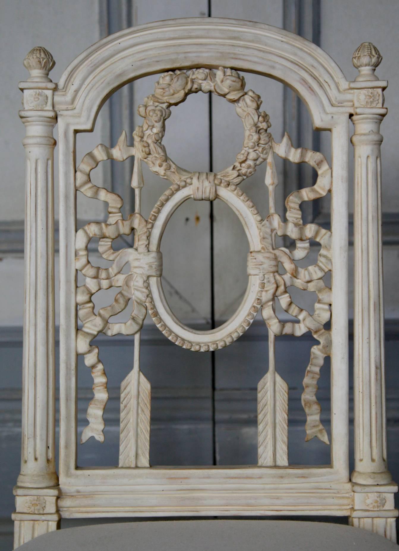 A Louis XVI style chair with intricate carving done by hand depicting classical renditions of arrows, ribbons and a floral wreath. Painted in antique white, slightly distressed and aged.