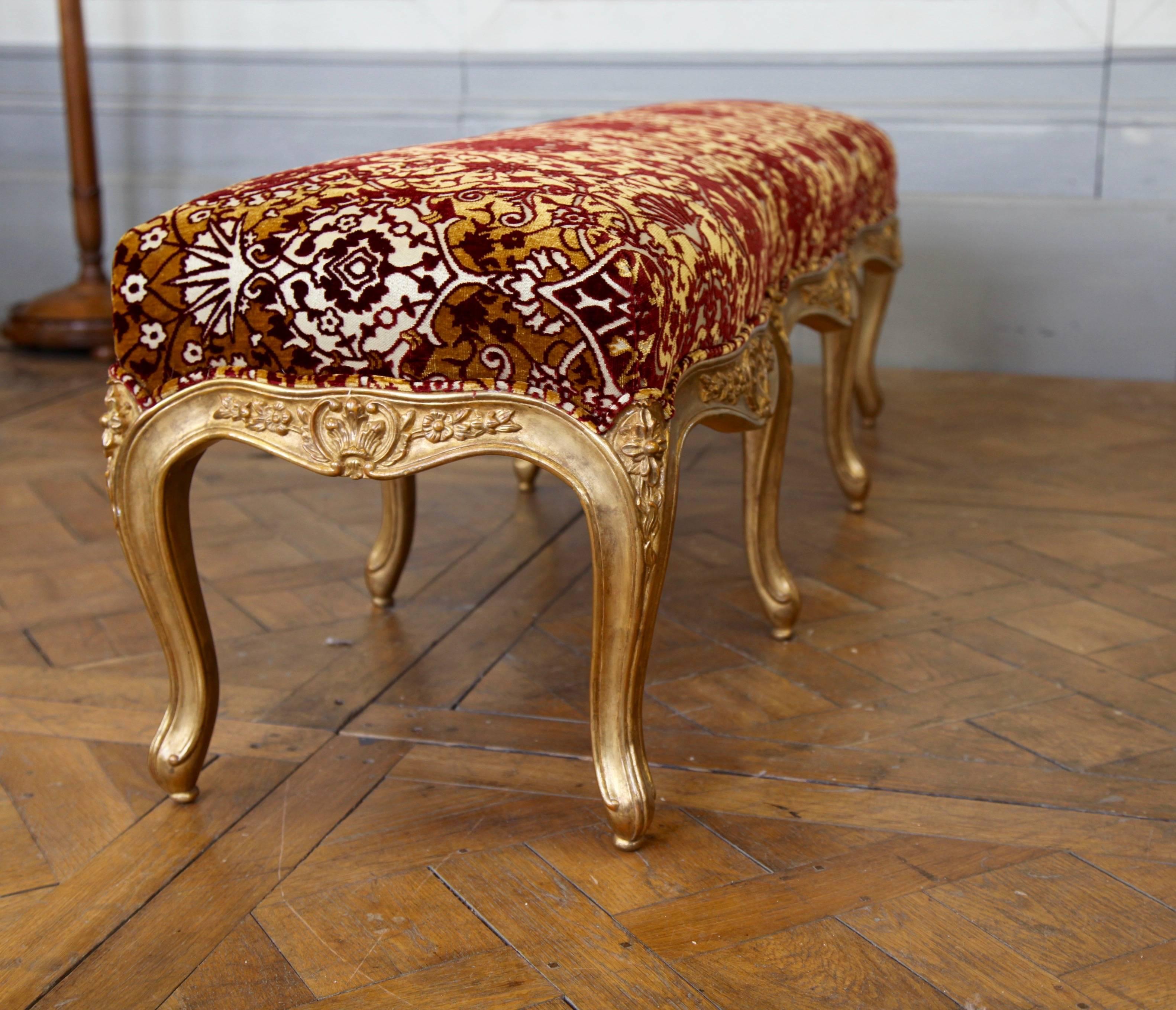 Louis XV style bench: Hand-carved by master craftsmen and hand finished in an antique gilded patina using traditional methods and materials. Upholstered in red and gold fabric as seen in the picture.