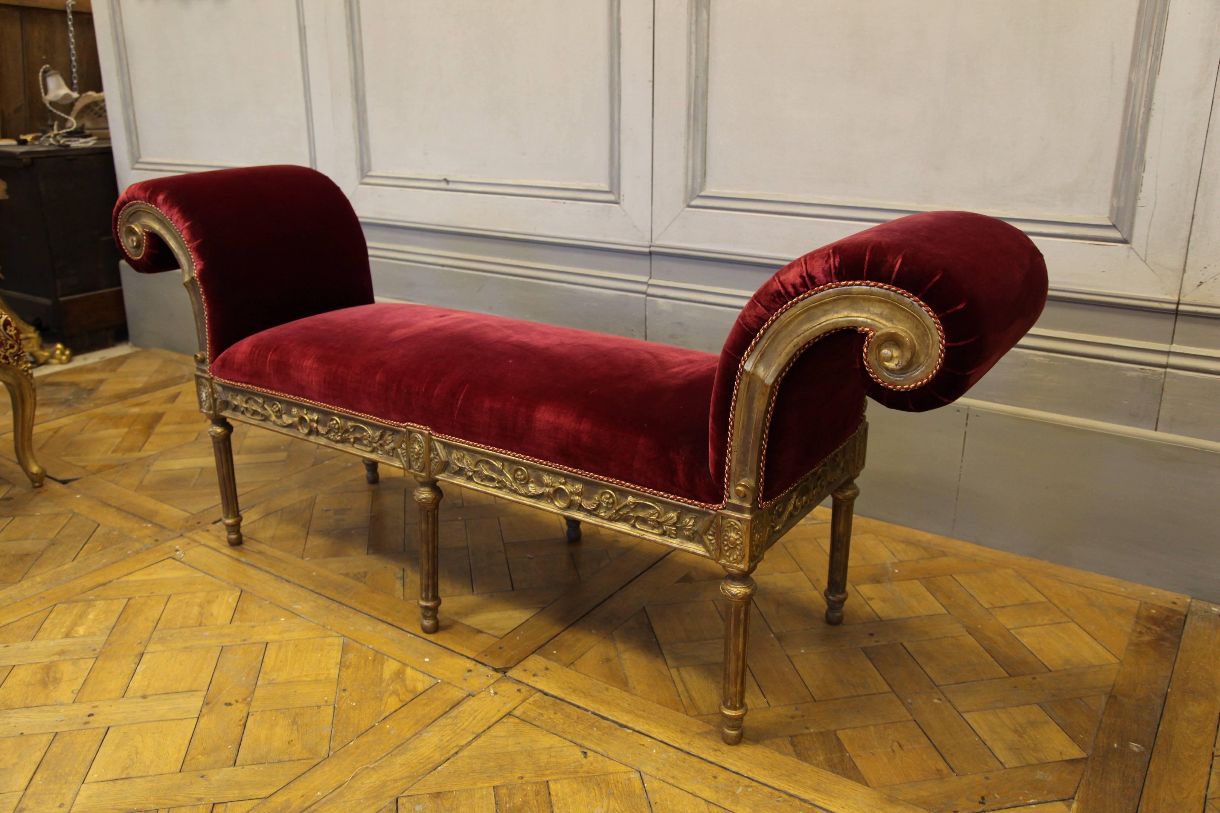 Louis XVI style giltwood banquette with scroll top arms: Hand-carved by master craftsmen and hand finished in an antique gilded patina using traditional methods and materials. Upholstered in a red velvet fabric with rope trim finish as seen in the