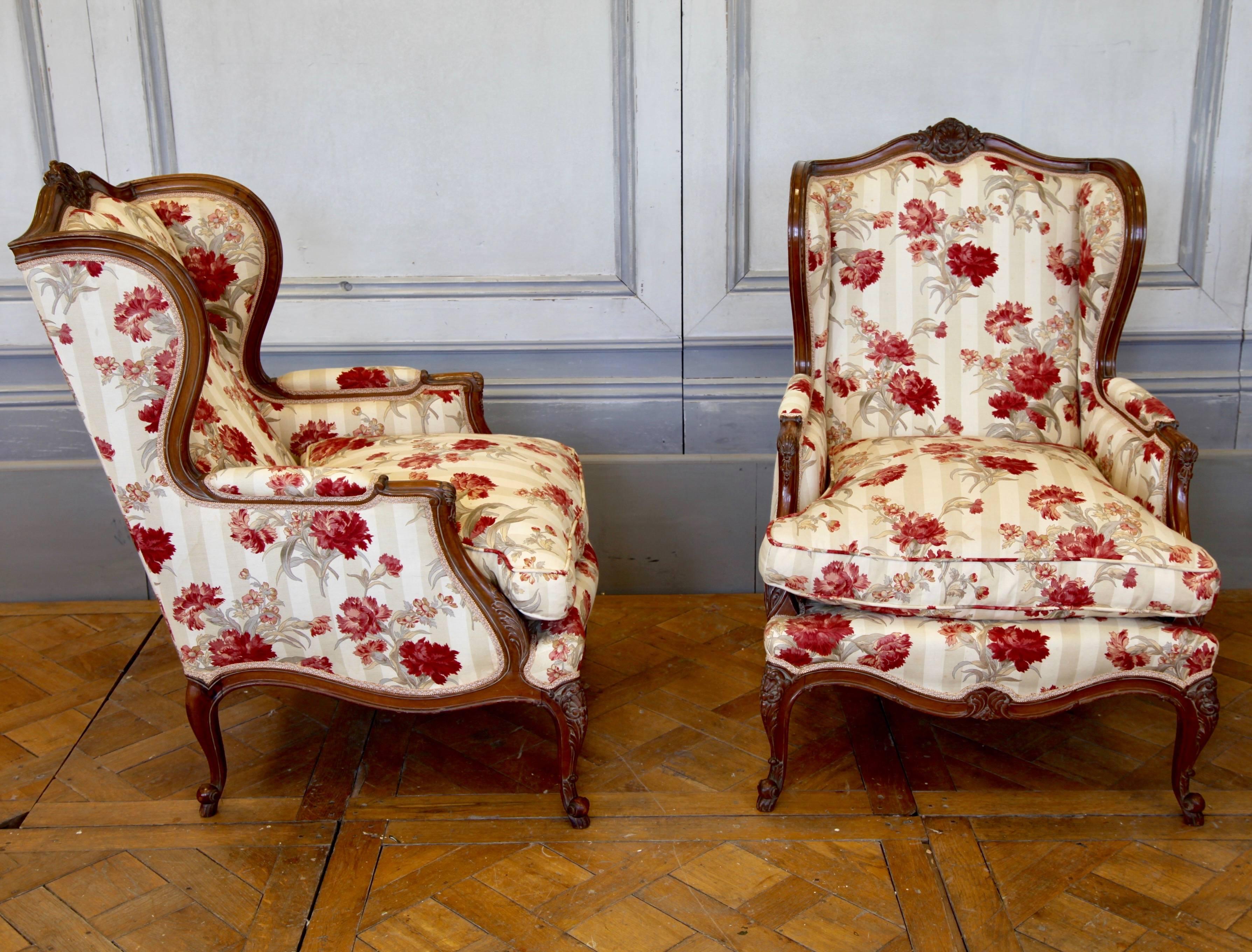 A charming pair of French antique Louis XV style bergere chairs from the early 20 century. hand-carved in walnut with scrolls and flourishes. Fully upholstered in a vintage fabric detailing red carnations.