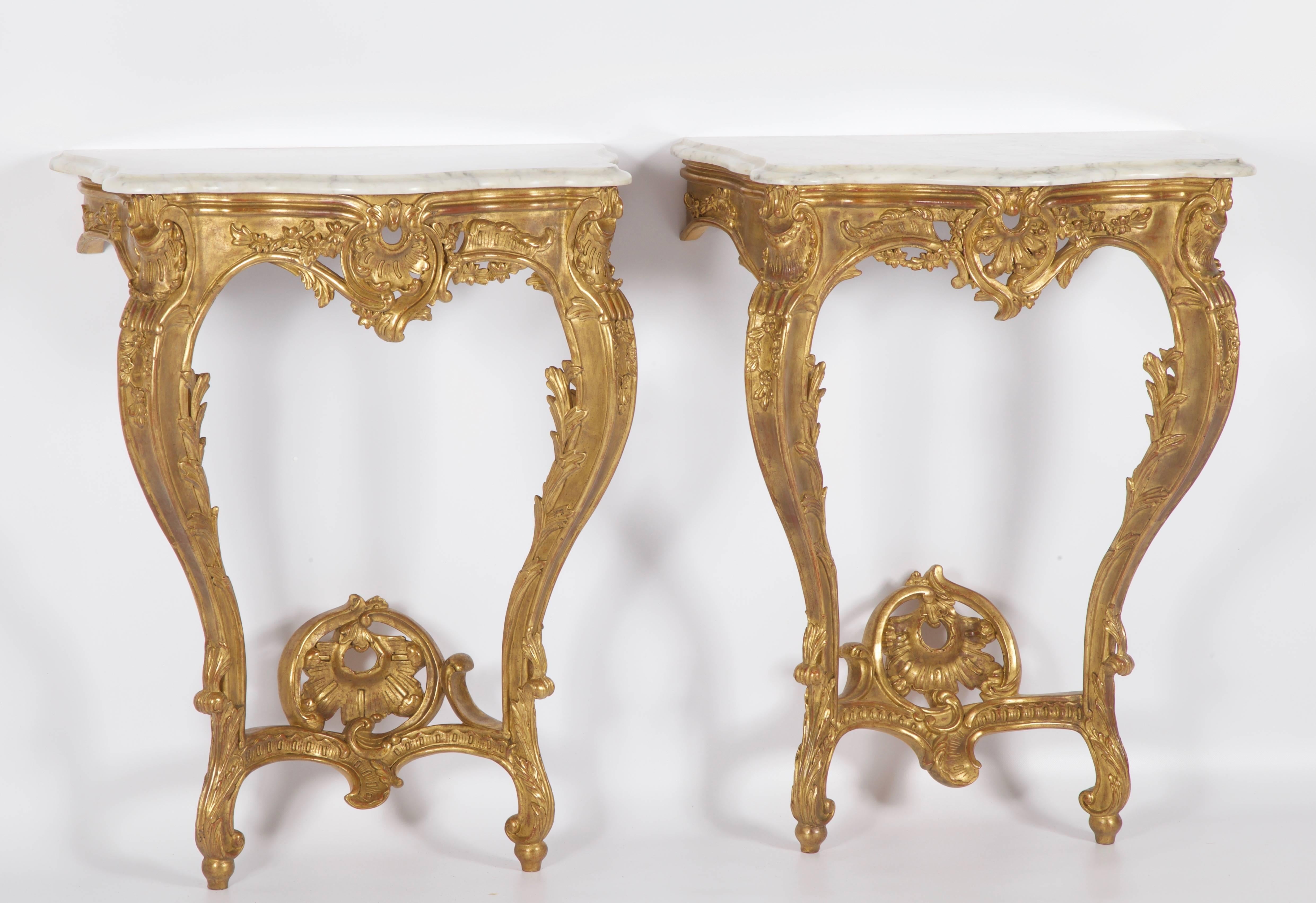 Louis XV style consoles hand-carved by master craftsmen and finished in a hand gilded patina slightly aged and distressed. Topped with Carrara marble which has been beveled, shaped and polished .
