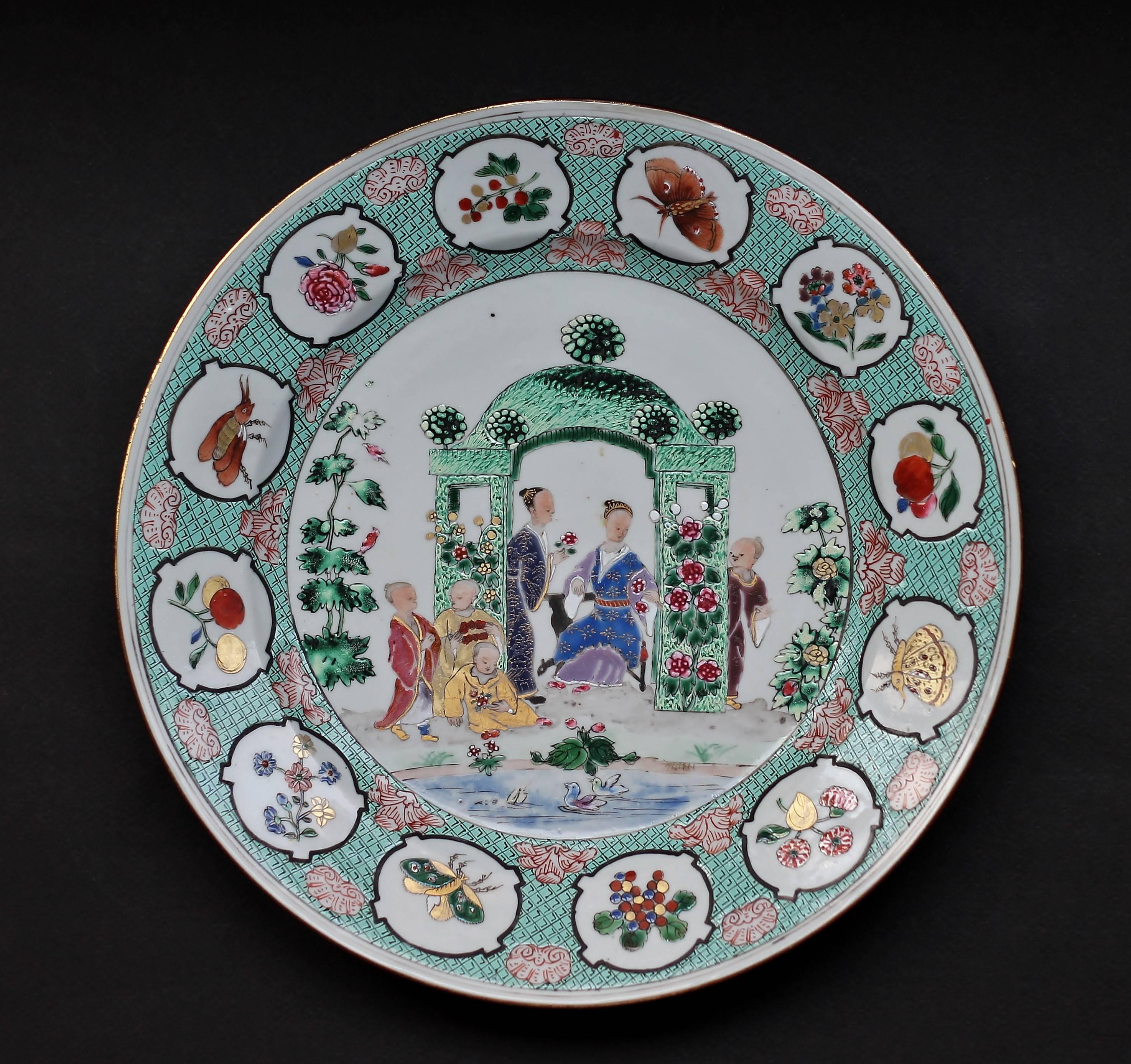 This enameled round dish with 'Arbor' design is decorated with a lady, servants and children by a pond, all dressed in Chinese costumes as perceived by the Europeans. Surrounding this scene are panels containing fruits, flowers and insects against a
