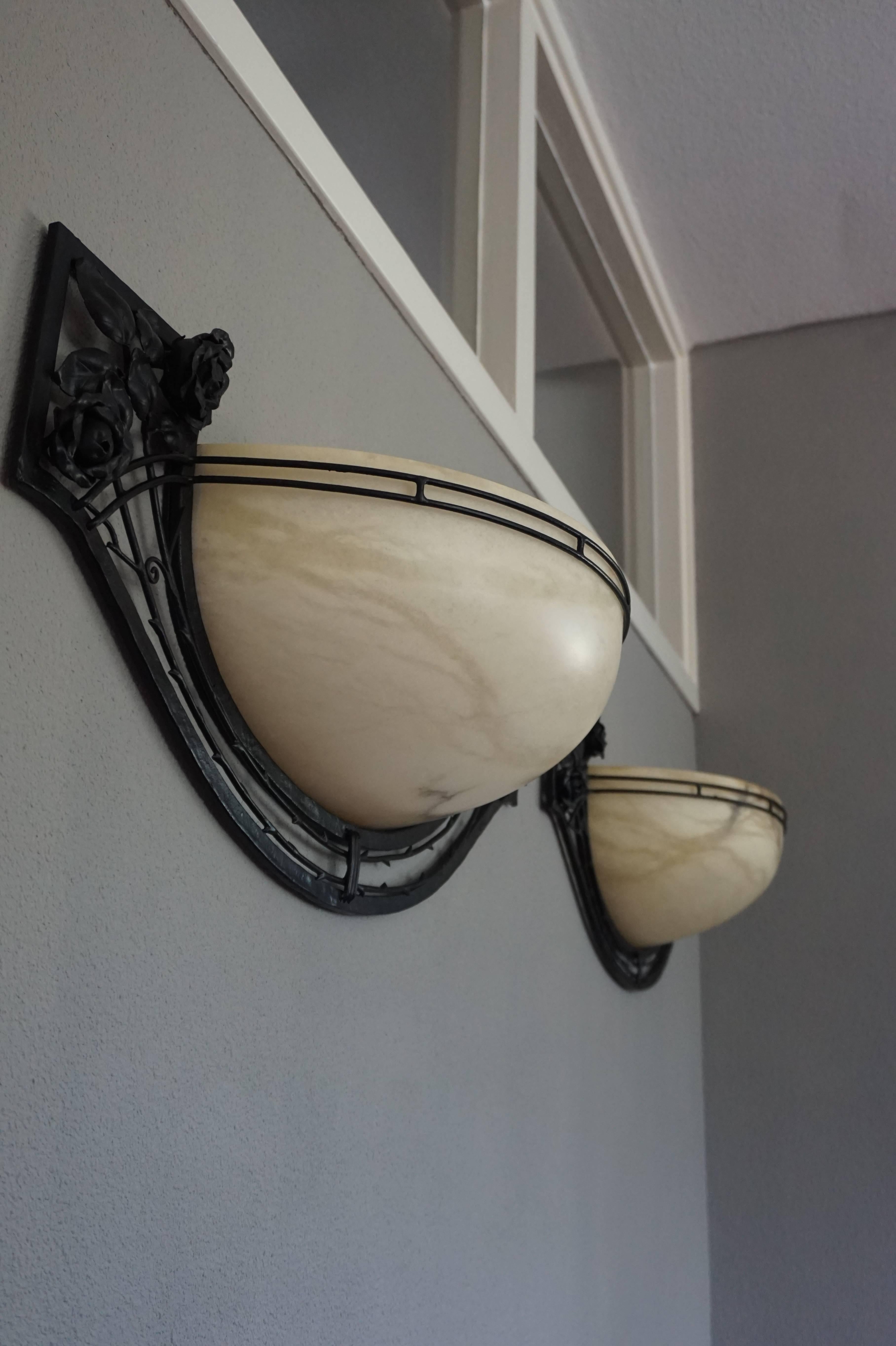 Impressive pair of sizable wall sconces with perfect alabaster shades.

This rare pair of Art Deco style sconces is beautiful both in design and execution. The combination of the blackened iron with the hand-crafted rose motifs and the creamy white,
