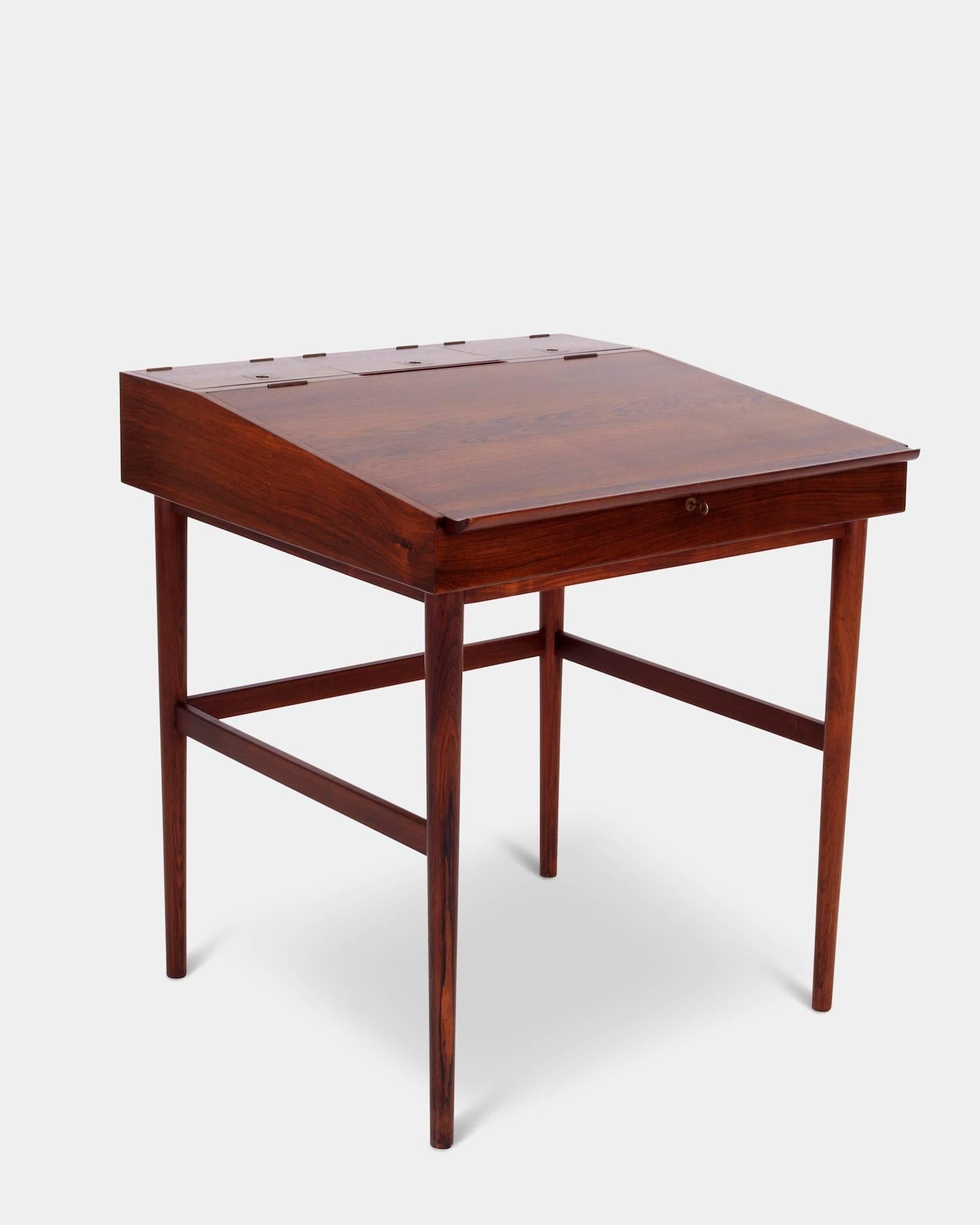 Model NV-40
A solid and veneerd rosewood writing desk.
Tabletop with flaps covering one large and three small compartments for storage. 
Brass handles and fittings, key included. Round tapered legs with stretchers 
Manufactured by Niels Vodder.